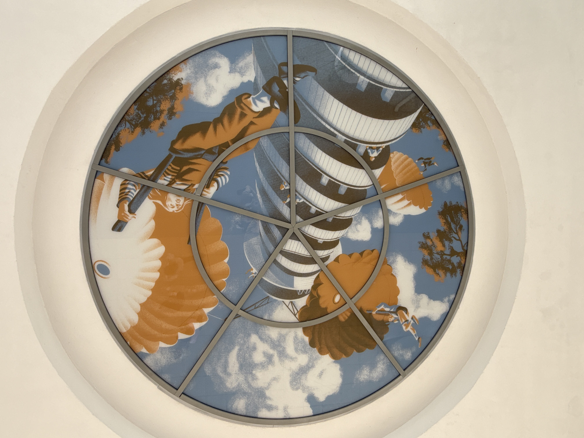 This is a circular stained-glass window depicting a stylized scene of paratroopers descending. The paratroopers are wearing orange jumpsuits and are descending under large, orange parachutes against a blue sky with white clouds. Below them are striped, cylindrical structures that appear to be buildings or towers. Trees with orange foliage are also visible in the background. The window is framed by a white, curved architectural element.