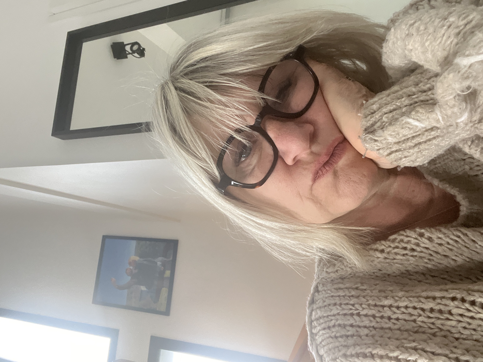 The image depicts an individual, appearing female with blonde hair, situated indoors in Chambœuf, France. The subject's upper torso and head are visible, angled slightly upwards, with her head resting on her right hand which supports her chin. Her lips are slightly pouted, and her eyes, observed behind dark, thick-rimmed glasses, appear partially closed or downcast. She is dressed in a light-colored, chunky knit sweater. The background features a white wall. To the upper left, a black-framed photograph is mounted, showing two figures outdoors with one arm raised against a blue sky. A portion of a window frame is visible below this picture on the left side. To the upper right, a large, black-framed mirror reflects a white ceiling and a black, ceiling-mounted light fixture.
