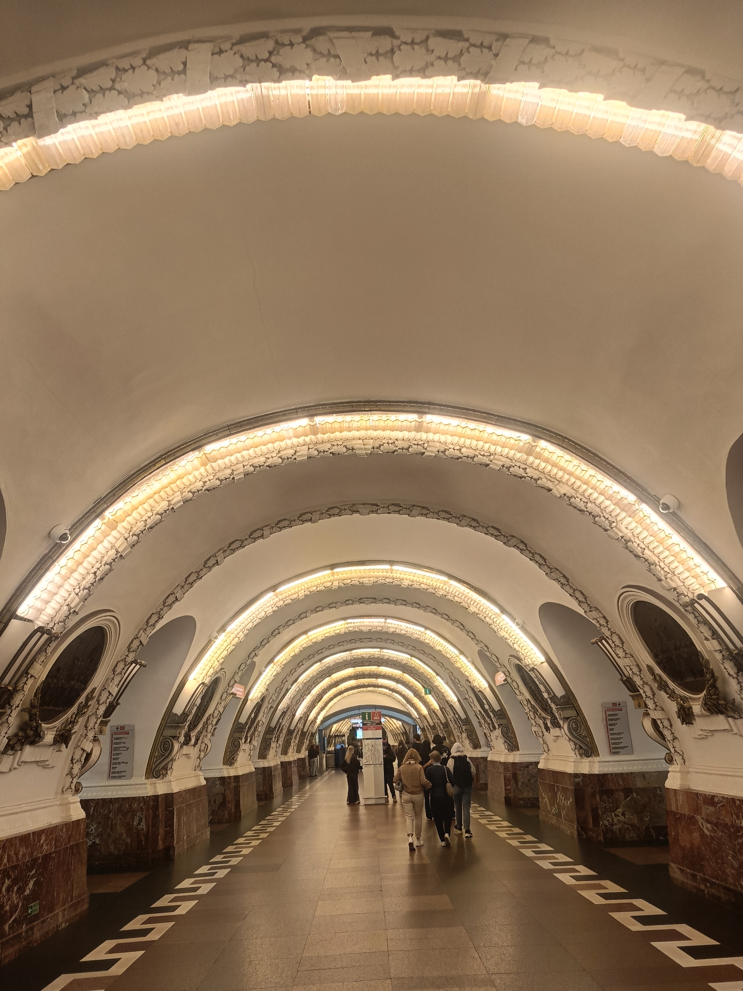 The image depicts a hallway with an arched ceiling in a subway station in Saint Petersburg, Russia. The ceiling is white, and the arches are decorated with ornate lighting and molding. The walls are white with marble panels at the bottom. The floor is tiled with a geometric pattern. People walk through the hallway. The lighting is dim and warm. The scene appears to be in a public transportation area, possibly a metro station.