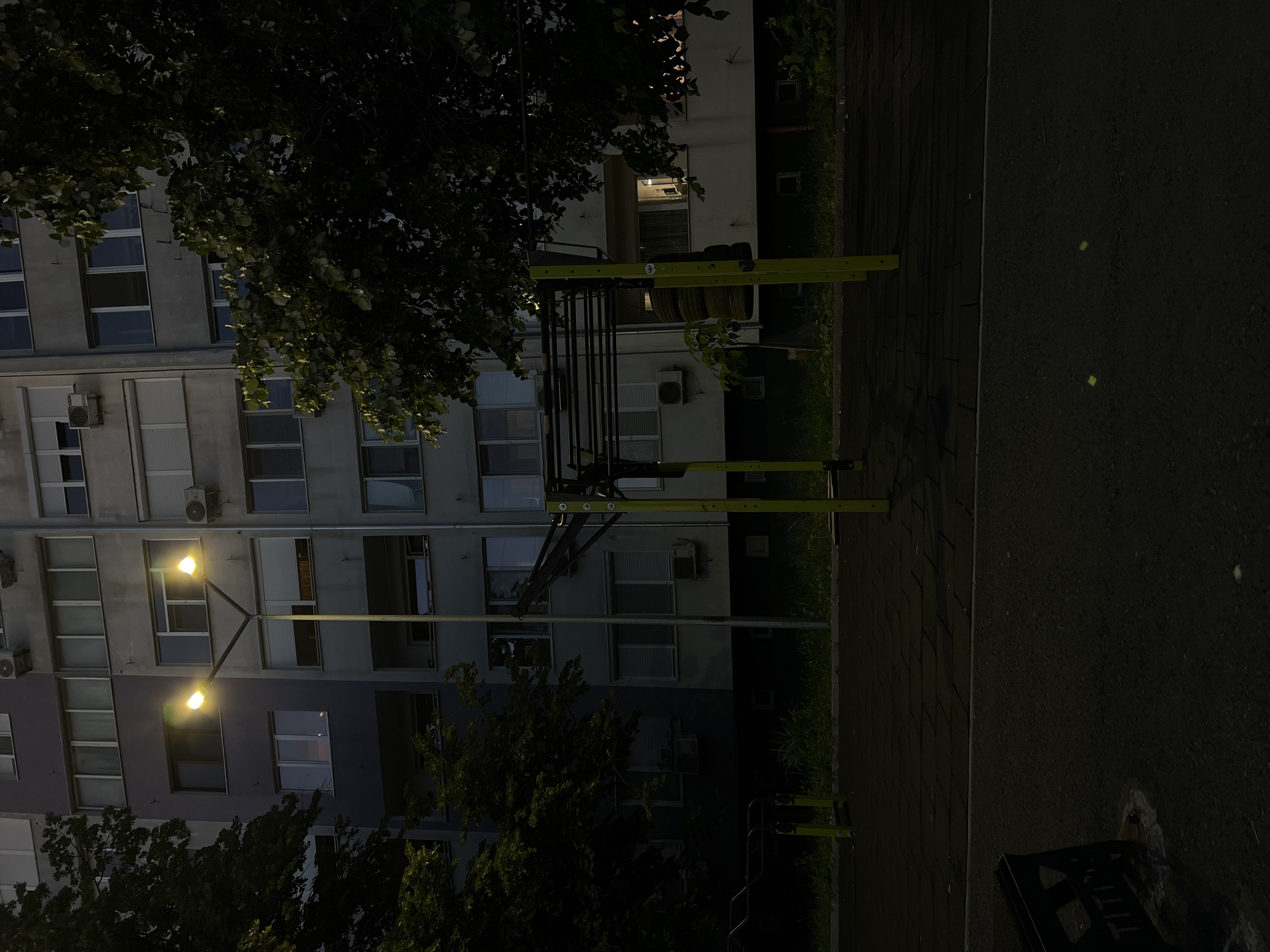The image depicts an outdoor exercise area next to a residential building in Burgas, Bulgaria, at dusk or night. A streetlamp with two lights illuminates the scene, casting a glow on the building's facade, which features numerous windows, balconies, and air conditioning units. To the right of the streetlamp is a jungle gym-like structure with yellow supports and black bars, used for calisthenics. Stacked tires hang from a horizontal bar on the structure. In the foreground, a dark, textured surface, possibly rubber matting or asphalt, serves as the ground cover for the exercise area. The left side of the image is partially obscured by the dark foliage of trees. The building appears to be multi-story, typical of urban residential blocks. No people are visible in the frame.
