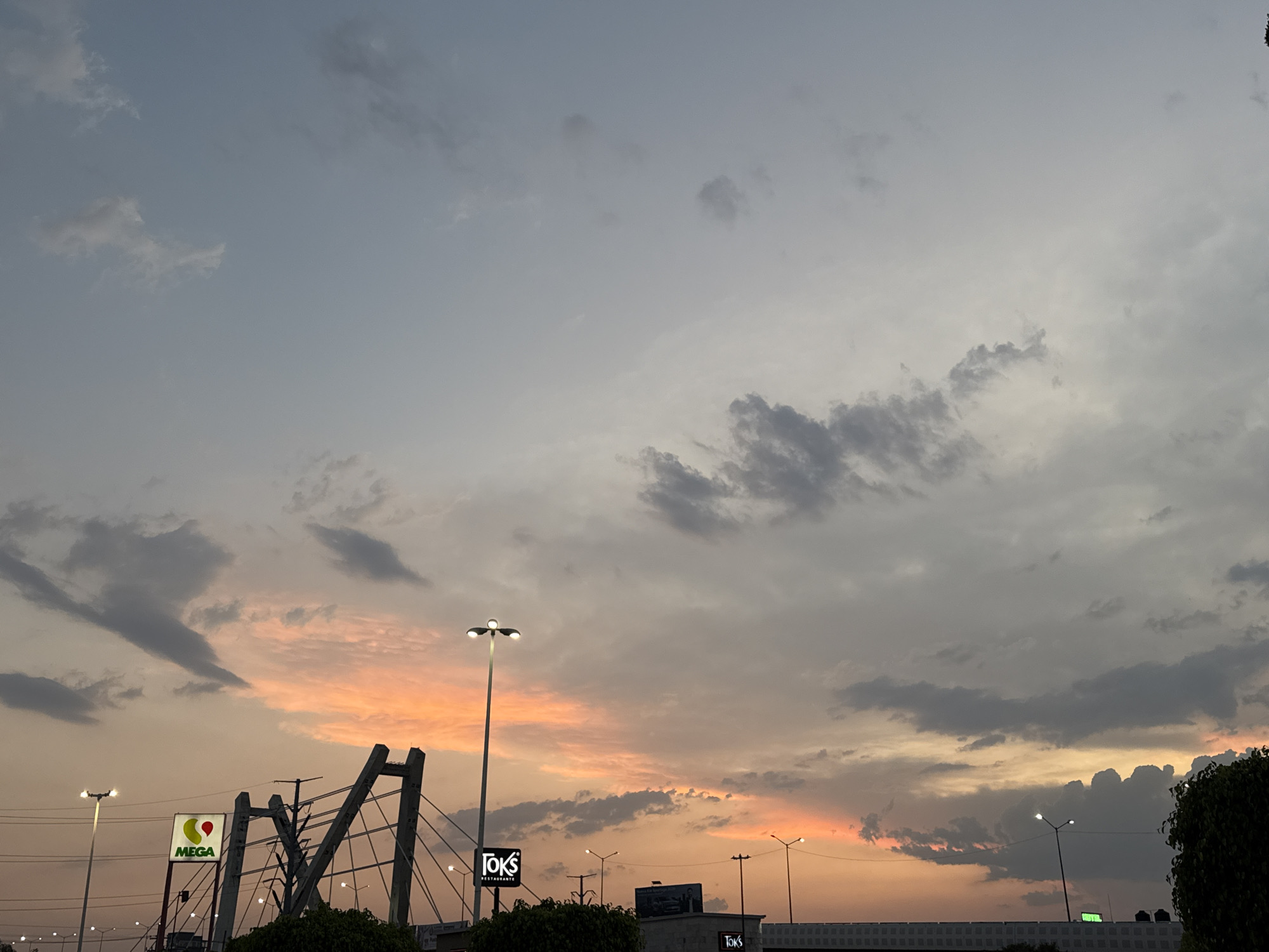 The image shows a twilight scene in the city of Puebla, Mexico. The sky is filled with clouds, transitioning from light gray at the top to orange and pink near the horizon. In the foreground, there are structures and commercial signs. A "MEGA" supermarket sign is visible on the left, alongside an arch structure. A "TOKS" restaurant sign is also present. Streetlights are scattered throughout the scene. The lower portion of the image shows vegetation and buildings. There are no people visible.