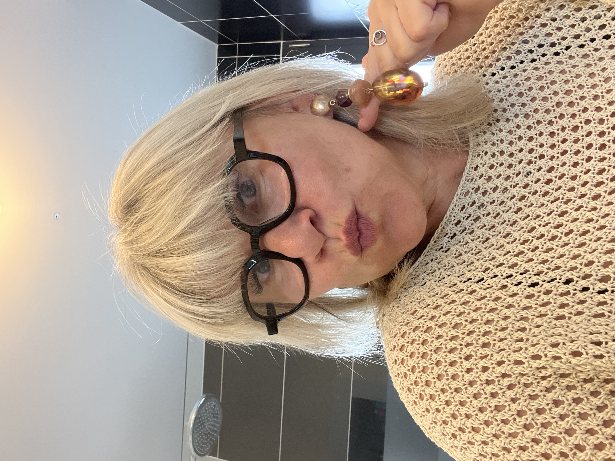 The image shows a woman with short, blonde hair and black-rimmed glasses. She is wearing a beige, crocheted top and large, beaded earrings. One of her hands is visible, adorned with a ring. The background appears to be a bathroom, with black and grey tiles and a showerhead. The location is Chambœuf, France.