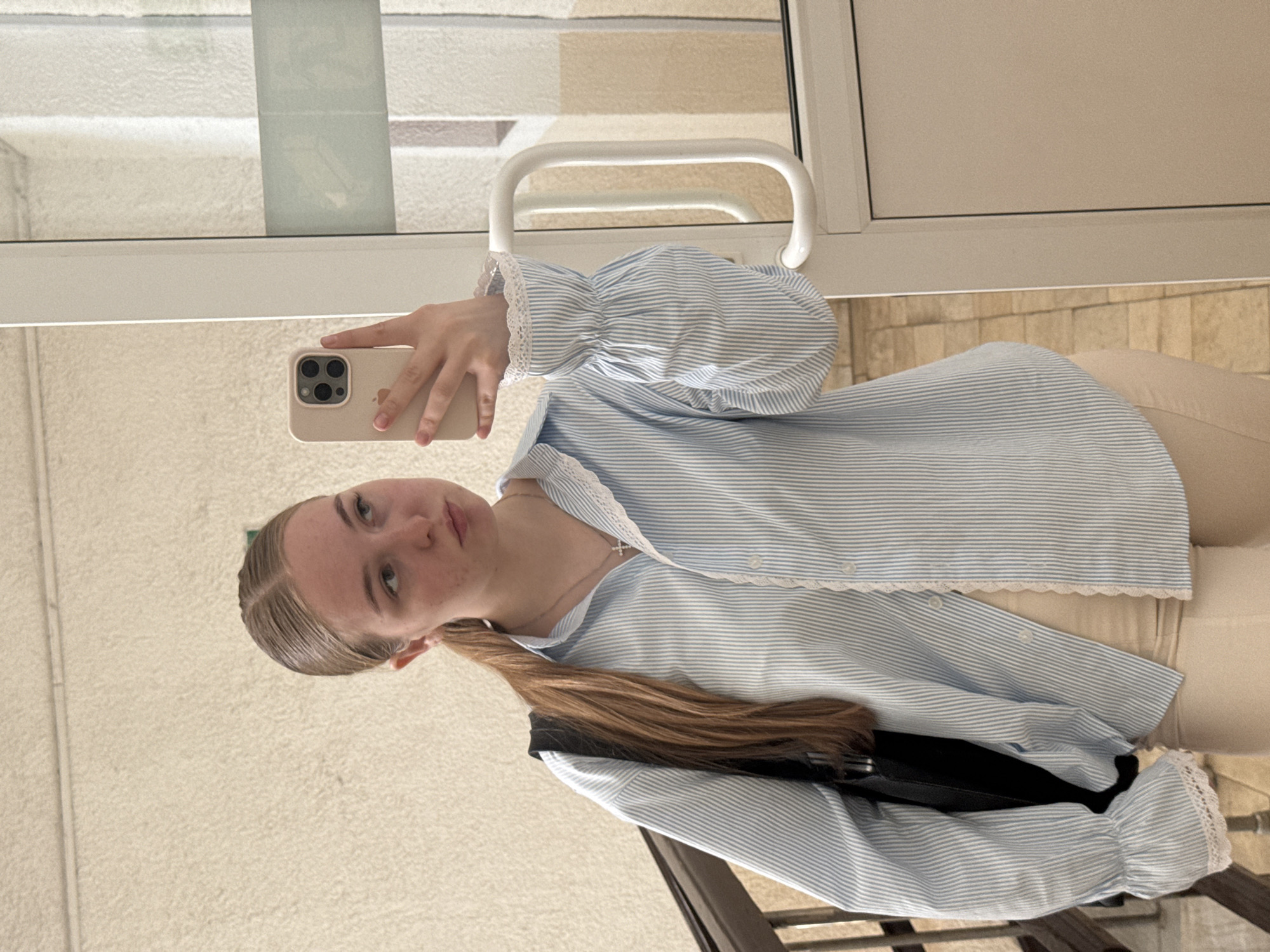 A young woman with light hair pulled back in a ponytail takes a selfie indoors near a window. She is wearing a blue and white striped button-down shirt with lace trim and light-colored pants. She holds a smartphone in her right hand, and a black bag is slung over her shoulder. The location is possibly in Perm, Russia, based on the user prompt. The environment appears to be a building's interior, indicated by a textured wall, a window frame, and a door handle.