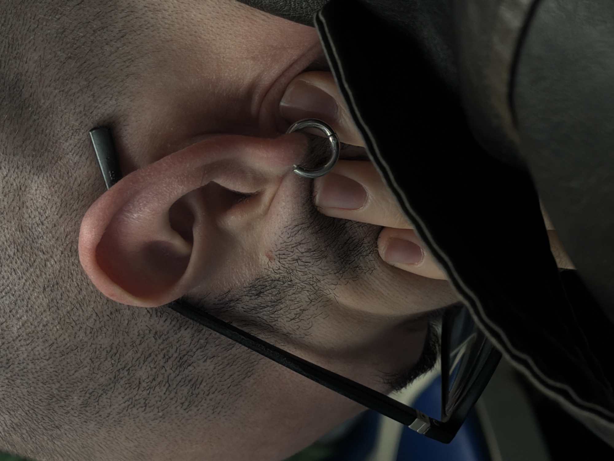 A close-up, angled view of a man's left ear with a silver hoop earring and a black straight post earring above the ear. The man is wearing black-rimmed eyeglasses. The person's fingertips are visible. A black jacket is nearby.
The location is unknown.
