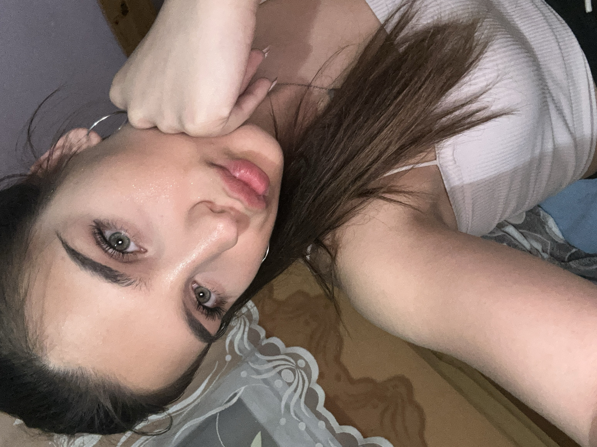 A close-up, overhead, selfie-style photograph captures a young woman from the chest up. She has light brown hair, fair skin, and light green eyes. Her right hand is resting on her chin, and she is wearing a light pink ribbed tank top and a silver hoop earring in her left ear. The background is out of focus, but appears to be an indoor setting with a patterned fabric behind her, possibly a bedspread or curtain, and a wooden surface to the right. There are no identifiable landmarks or objects that would indicate a specific location within the city of Shumen, Bulgaria.