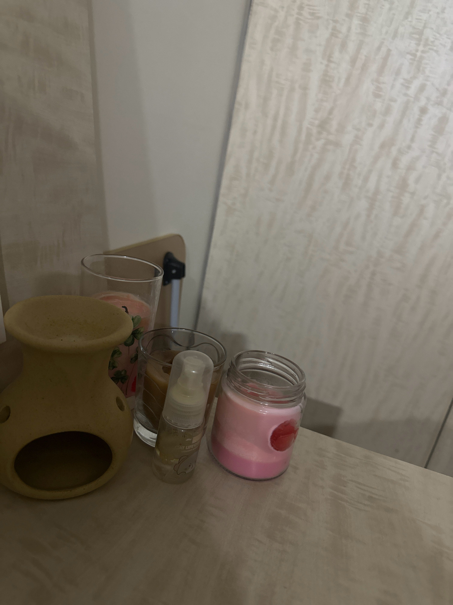 A beige ceramic oil burner sits on a textured wooden surface, with several candles and a small spray bottle beside it. To the left of the oil burner, a clear glass contains a pink candle with green clover patterns. Next to that, another glass holds a brown candle. In front of these, a clear spray bottle with a white cap and a light yellow label featuring a cartoon cat is visible. To the right, a pink candle in a glass jar with a red circular decal is present. In the background, a light-colored wall with a subtle pattern is partially visible. The scene is dimly lit, casting shadows.