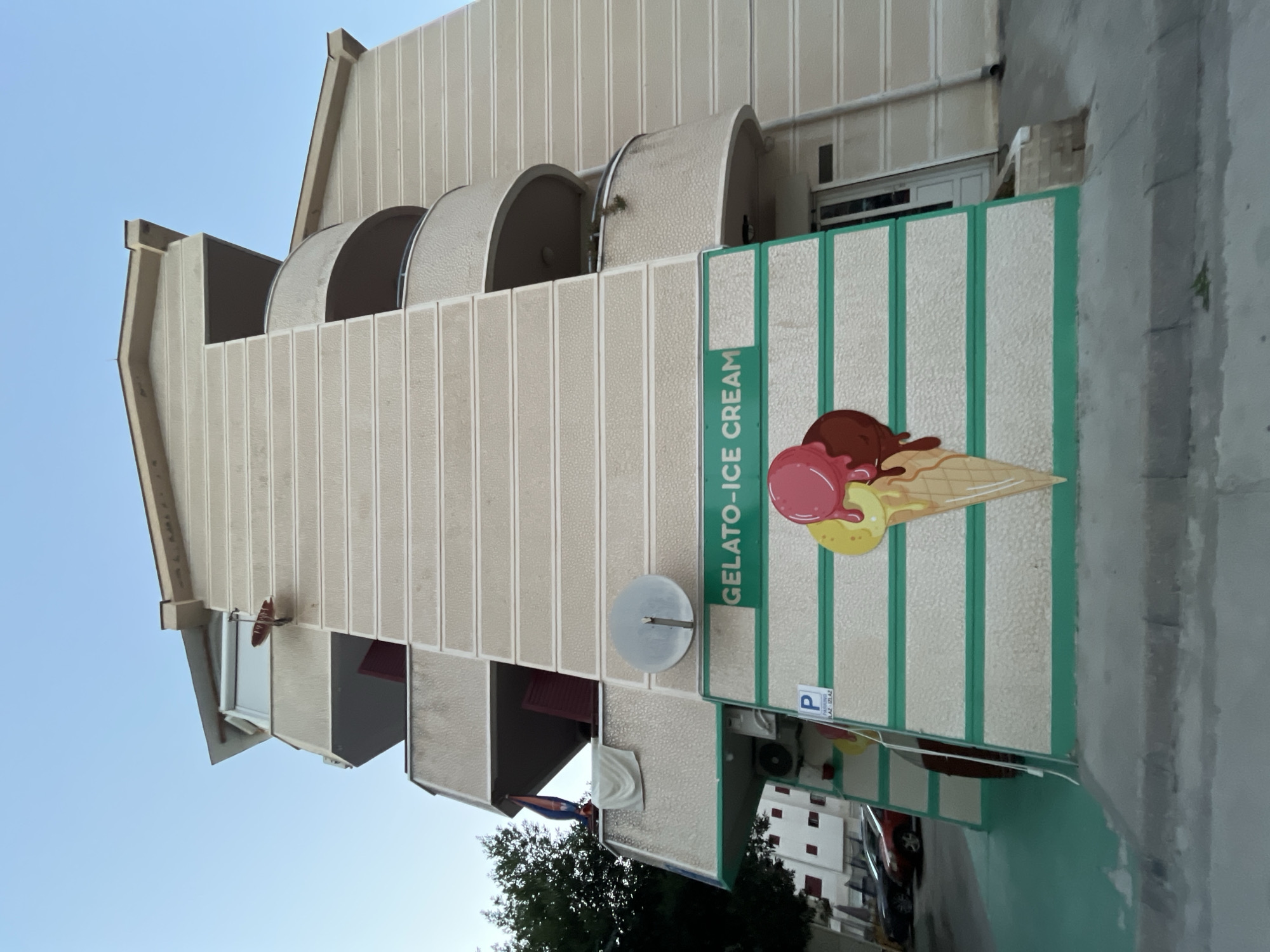 This low-angle, outdoor, wide shot captures the facade of a multi-story building in Budva, Montenegro, with a clear blue sky overhead. The building is light beige with horizontal textured lines and features several balconies with curved railings. The ground floor is decorated with a large mural of an ice cream cone with three scoops of different colors (pink, yellow, and brown) and the words "GELATO-ICE CREAM" in green lettering. Green stripes accent the lower section of the building's facade. To the left, a satellite dish is mounted on the building. Below, a dark green painted area extends outwards, and a portion of a street with parked cars, including a red sedan, is visible. A sign with a blue 'P' indicating parking is also present. The environment suggests a commercial or residential building in a warm climate.