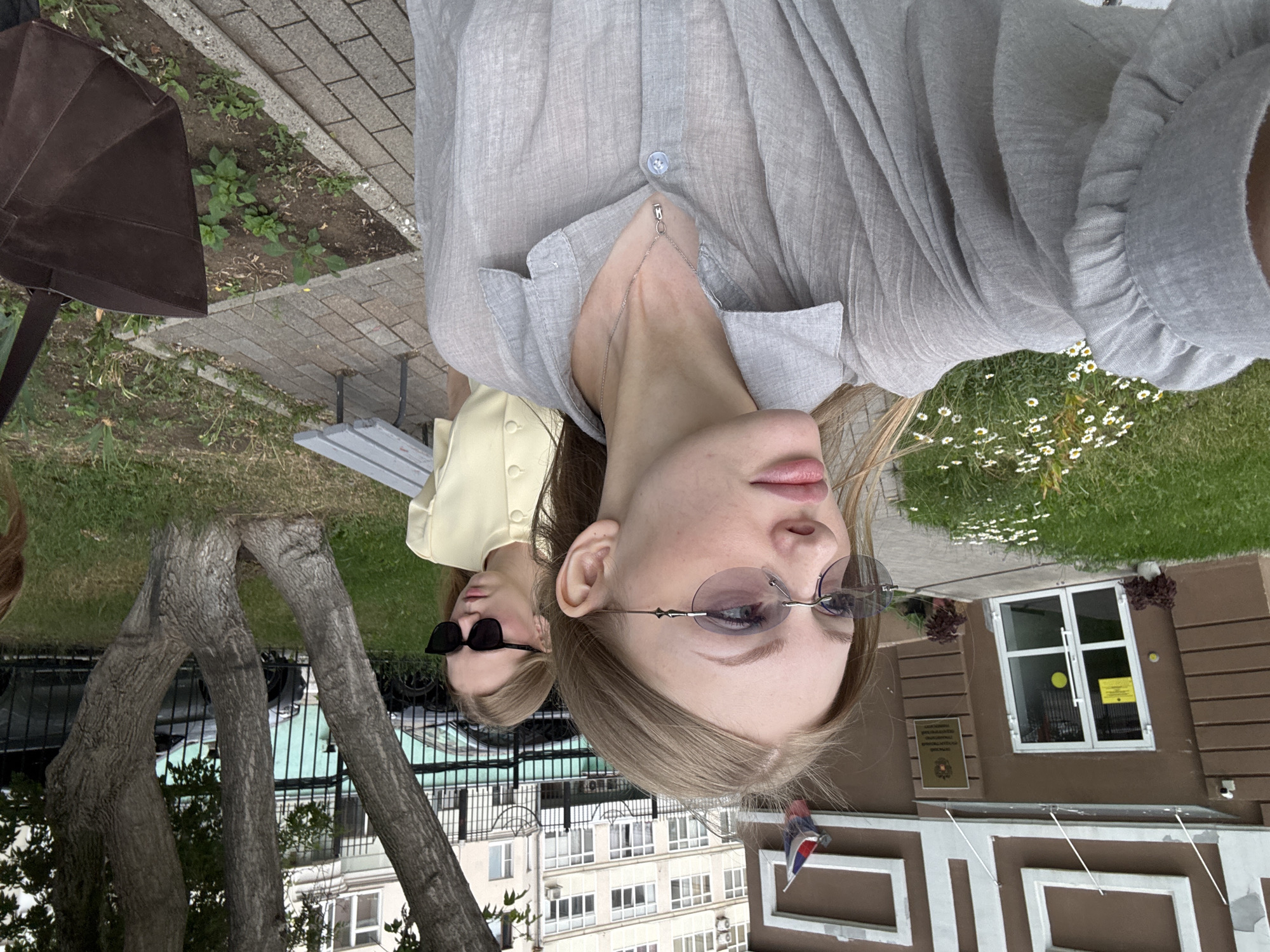 In Perm, Russia, two young women are captured in a selfie outside a building. The woman in the foreground, with blonde hair and sunglasses, is wearing a light grey, translucent blouse with a necklace. Behind her, another blonde woman in a light yellow top and sunglasses looks away from the camera. The building appears to be a government institution, indicated by the Russian flag and signage visible on its facade. The surrounding environment includes a paved walkway, a grassy area with small white flowers, trees, and parked cars in the background. A grey bench is situated near the building.