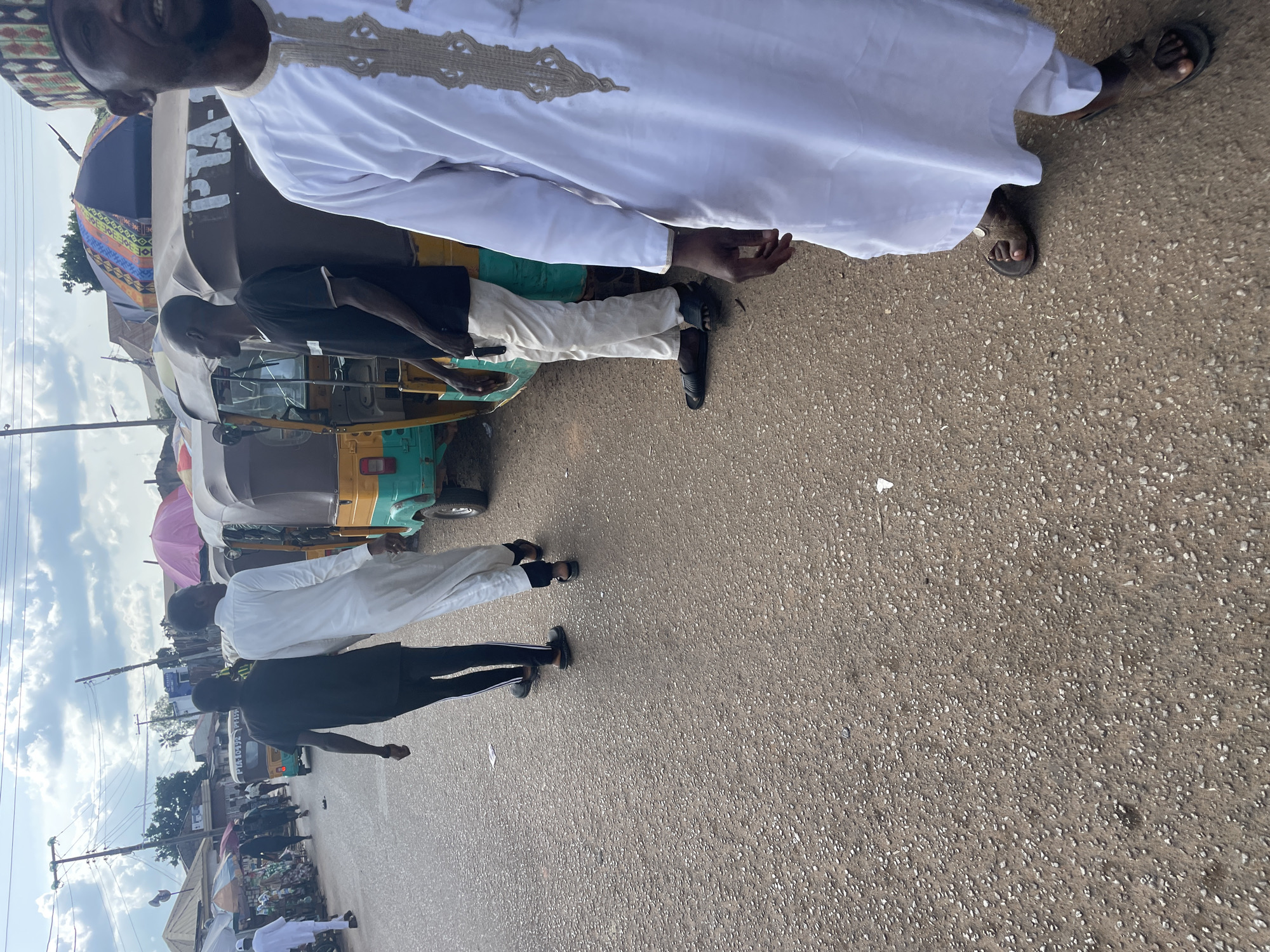 The image shows a street scene in Gura Top, Nigeria. In the foreground, a person wearing a white robe and sandals walks away from the camera, their face partially visible. To their left, another person in a black t-shirt and white pants with black stripes also walks away, holding sunglasses in their right hand. Behind them, several auto rickshaws, some with passengers, are parked or moving on the street. The background features stalls with colorful umbrellas, poles with overhead wires, and buildings. The ground is a rough, gravelly asphalt surface. The sky is partly cloudy.