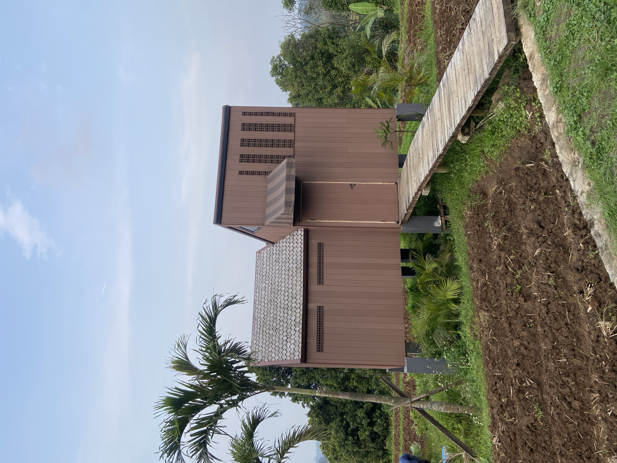 The image shows a two-story brown building with a tiled roof and a striped awning in Selotapak, Indonesia. A wooden walkway leads to the building, situated beside a field of tilled earth. A palm tree stands in the foreground, while a variety of trees are visible in the background. The sky is light blue with some cloud cover.