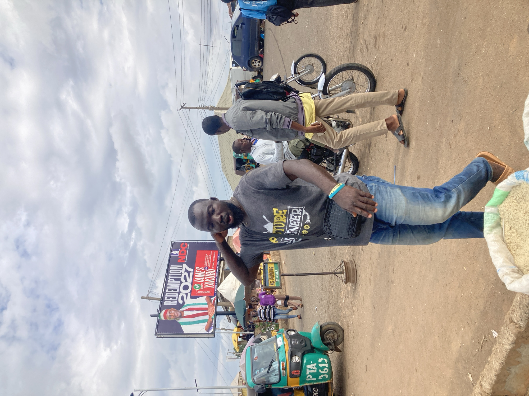 The image depicts a street scene in Gwong, Nigeria, under a cloudy sky. In the foreground, a man wearing a gray t-shirt and blue jeans stands with his right arm raised to his head, looking off to the side. He has a black crossbody bag slung over his shoulder. To his left, a green and yellow motorized rickshaw is parked. Behind him, a billboard displays a political advertisement for "REDEMPTION 2027 NDC" featuring a portrait of a man identified as "HON. JAMES YAKUBU." Several other individuals are visible in the background, including two women walking and two men on a motorcycle. A "NO PARKING BY ORDER" sign is also present. The ground is paved with dirt and gravel. In the lower right corner, a large white sack filled with what appears to be grain is partially visible.