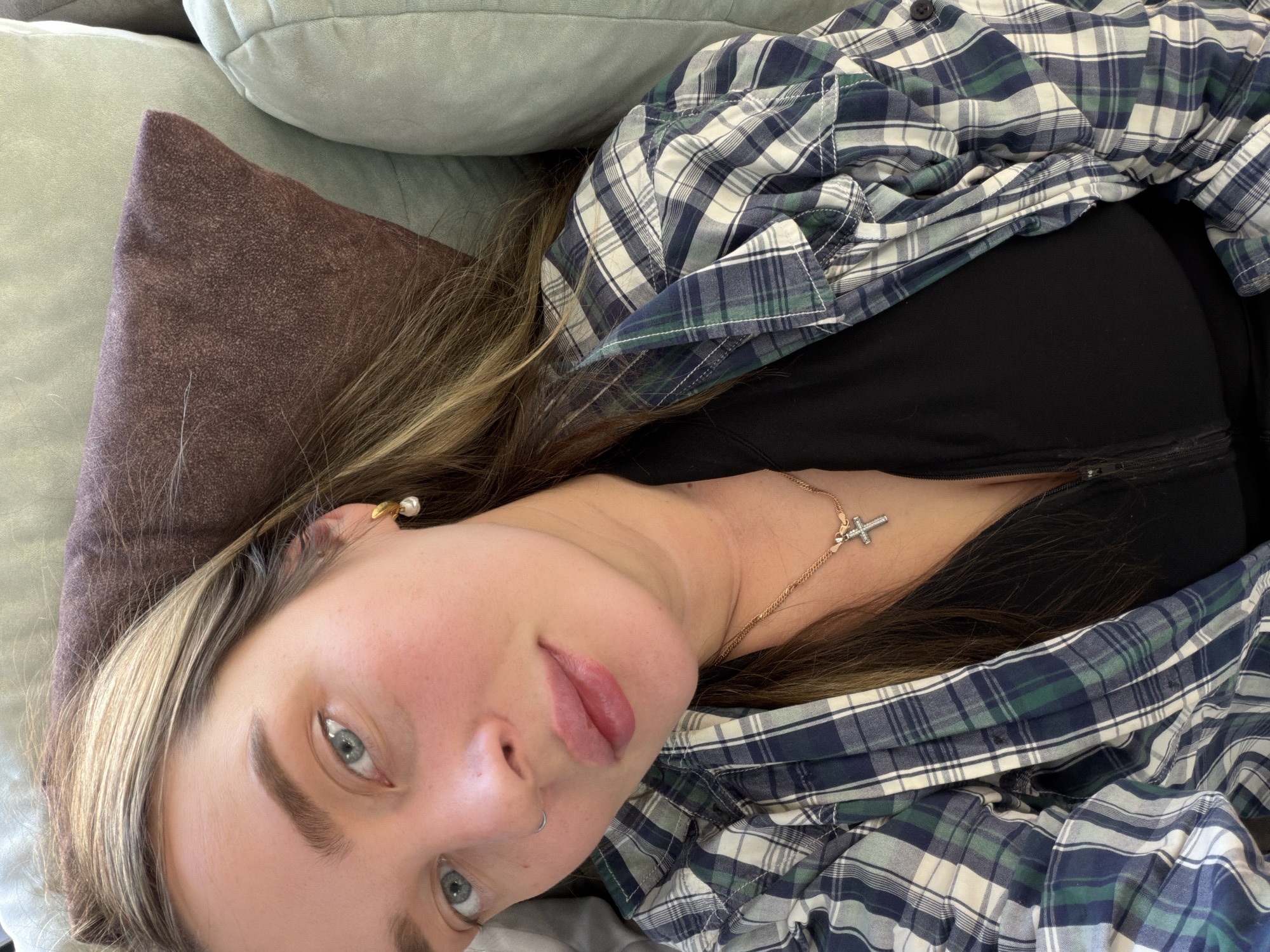 The image shows a woman lying on a light grey cushion. Her head rests on a brown pillow. She is wearing a black top and a blue and green plaid shirt. A gold necklace with a cross pendant is visible on her neck. She has light blue eyes, fair skin, and light brown hair with blonde highlights. She has a nose ring in her left nostril and an earring in her right ear. The background also includes a light green pillow. There is no indication of the location being Chaikovsky, Russia.