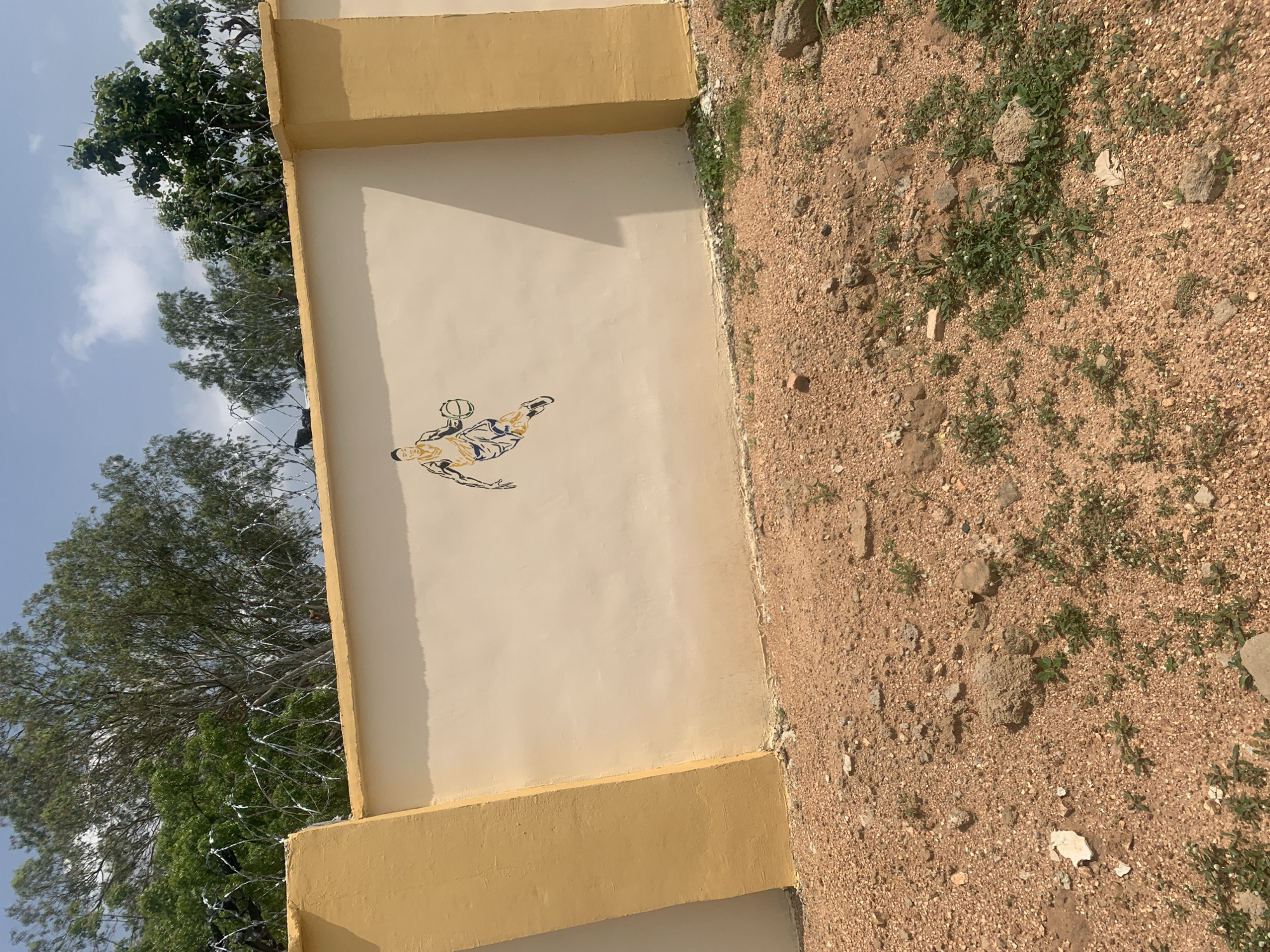 The image shows a section of an outdoor wall in Jalingo, Nigeria. The wall is painted a light cream color with yellow vertical support beams at intervals. A stencil-like mural of a basketball player in motion is painted on the cream section of the wall. The player is depicted in a blue and yellow uniform, dribbling a basketball. The top of the wall is topped with barbed wire. Behind the wall, several trees with green foliage are visible, along with a blue sky with scattered white clouds. The ground in front of the wall is a reddish-brown gravelly terrain with sparse green vegetation.