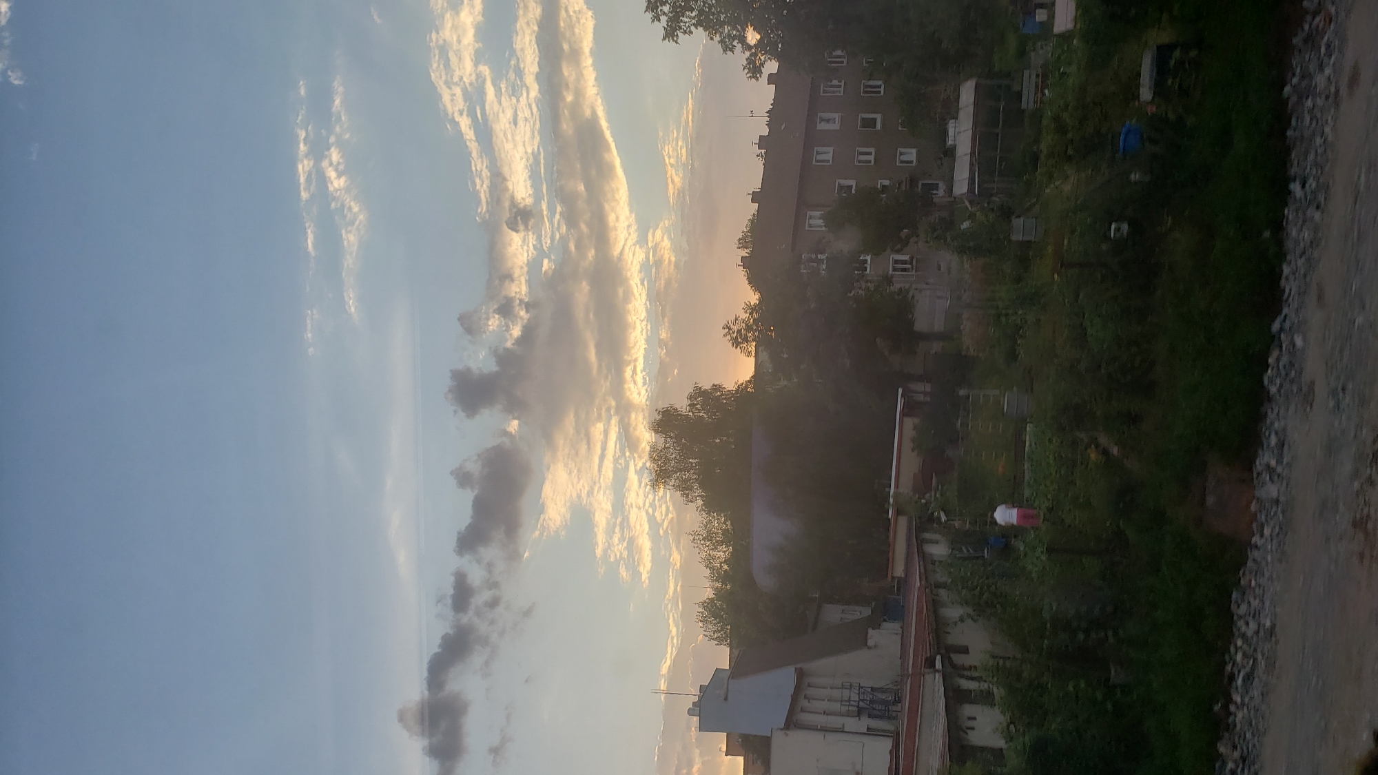 The image depicts a residential area in Poprad, Slovakia, at dusk. The sky is a pale blue with scattered clouds tinged with orange and yellow, indicating the setting sun. In the foreground, dense green foliage and vegetation line a gravelly embankment. Behind the vegetation, several buildings are visible.
On the left, a light-colored building with a blue section stands prominently, featuring visible windows and an external metal staircase. Adjacent to it, a single-story structure with a reddish roof is partially obscured by trees. Further to the right, a multi-story apartment building with numerous windows is visible, its facade a muted brown. A greenhouse structure is also present in the middle ground, alongside other smaller utility buildings.
In the middle of the garden area, a lone person wearing a white top and pink trousers is standing, facing away from the camera. The overall scene suggests a quiet evening in a suburban or semi-urban setting.
