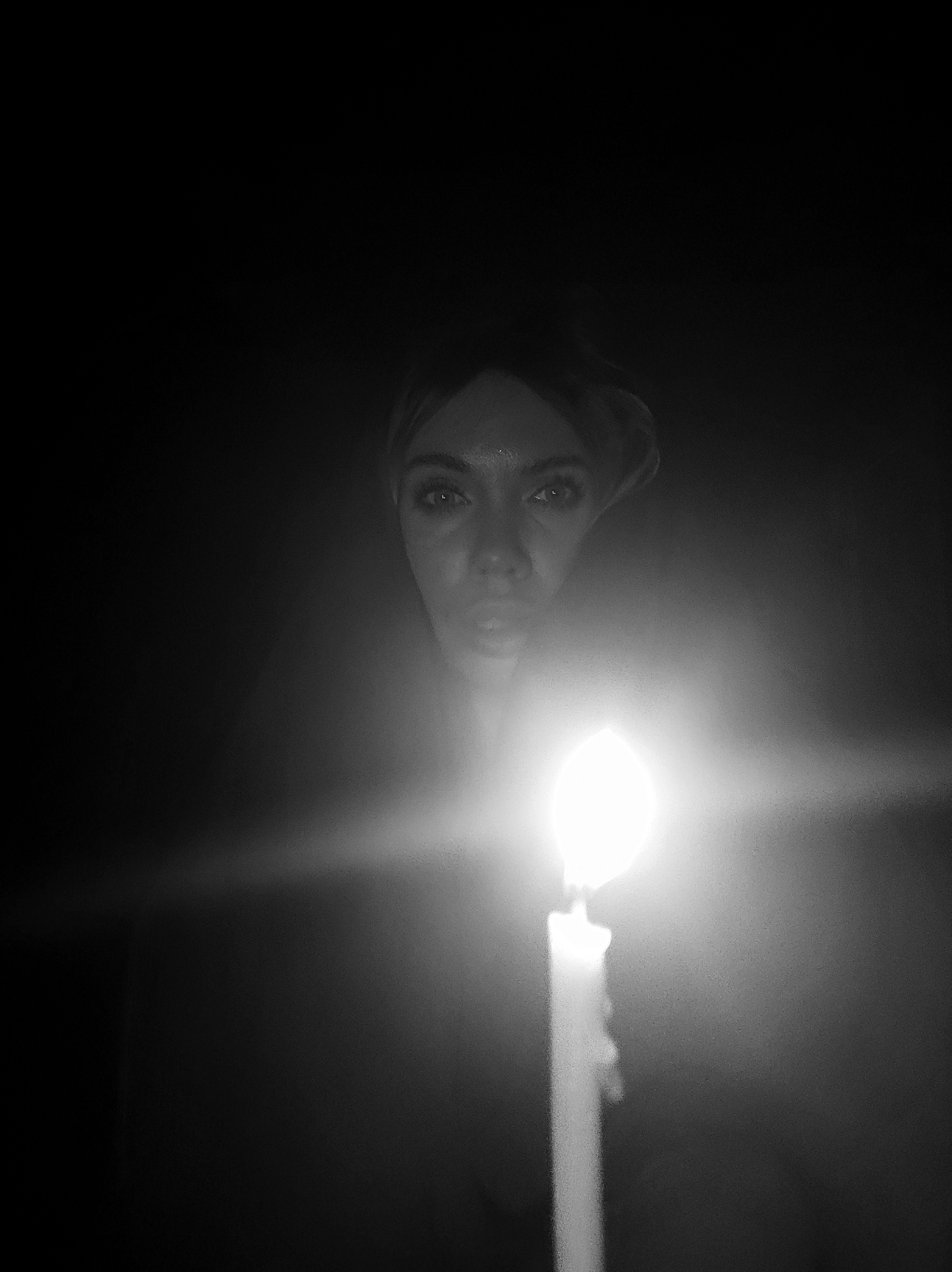 A black and white image features a person holding a lit candle in the dark. The light from the candle illuminates the person's face, revealing their eyes and mouth. The environment is otherwise indistinguishable due to the lack of light. No information suggests a location in Votkinsk, Russia.