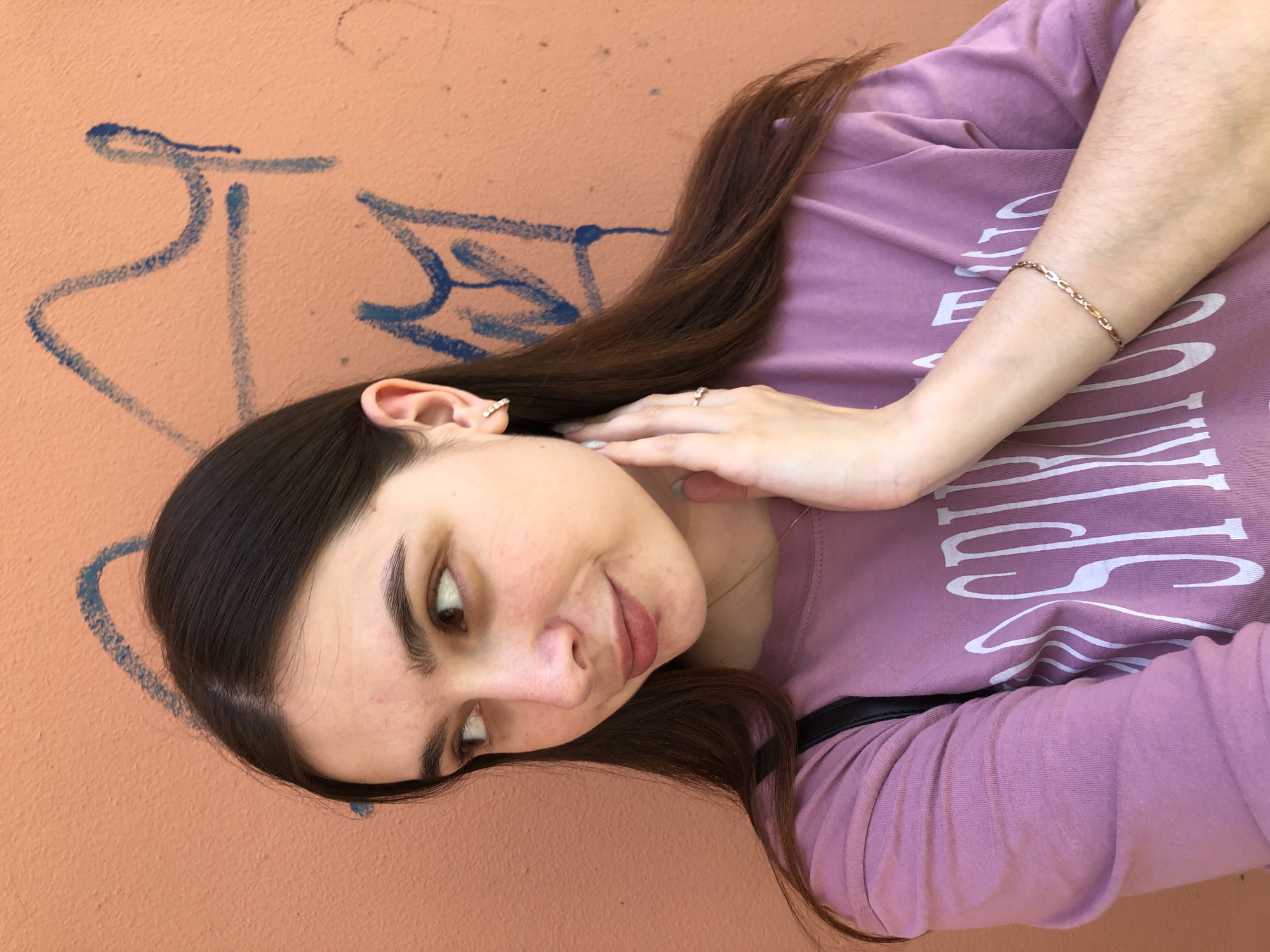 A young woman stands against an orange wall with blue graffiti. She has brown hair, brown eyes, and fair skin, and she is wearing a long-sleeved, lavender t-shirt and gold jewelry, including a bracelet, rings, and an earring. Her right hand touches her face. The location is in Chaikovsky, Russia.