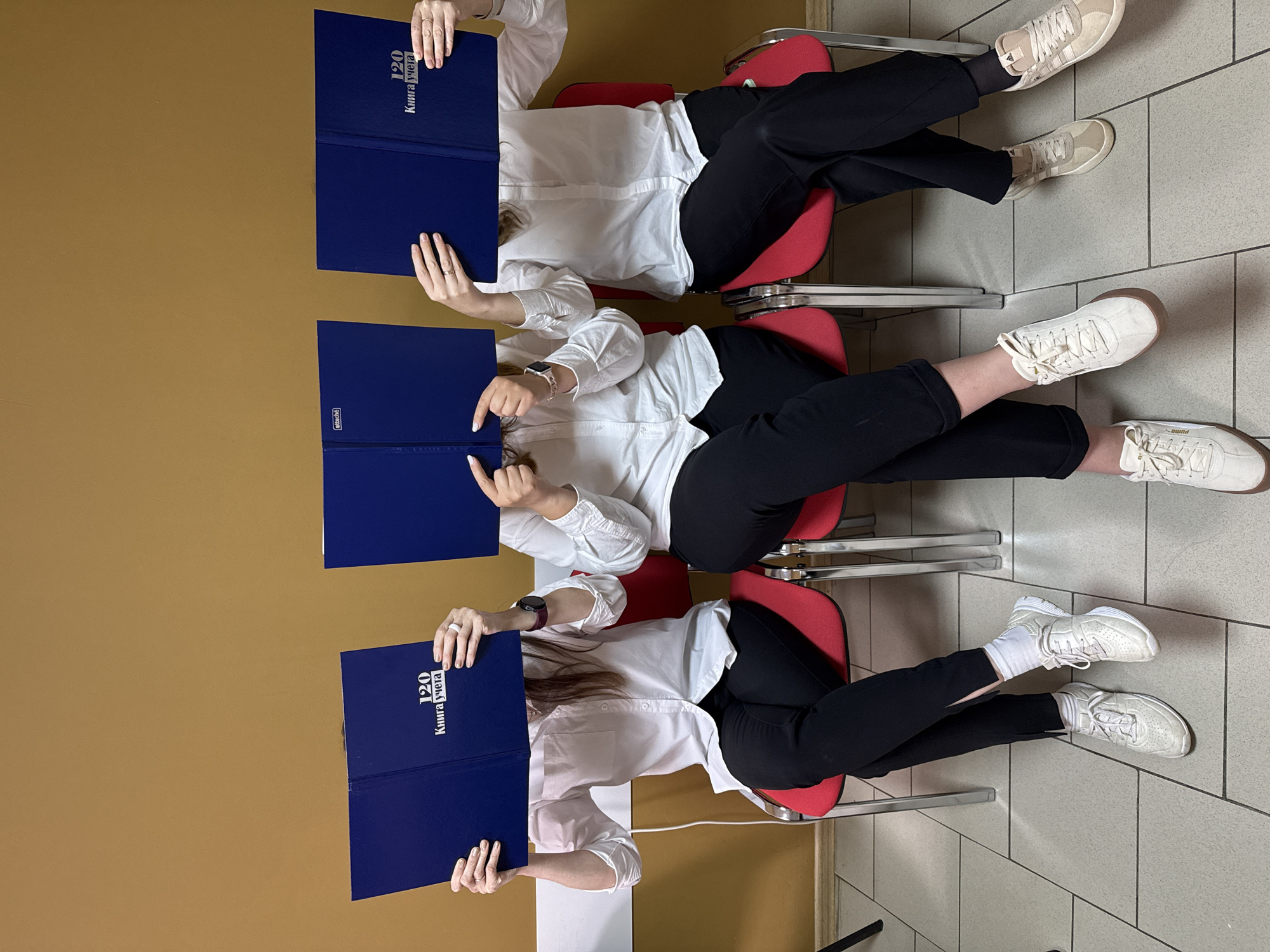 Three individuals sit in a row of red chairs in a room with a brown wall on the left and a tiled floor on the right. Each person wears a white shirt and black pants, and each holds a blue folder in front of their face, obscuring it from view. The folder has text in Cyrillic that reads "120 Kniga ucheta". All three individuals wear white sneakers. The image is rotated 90 degrees clockwise. The location is Chaikovsky, Russia.