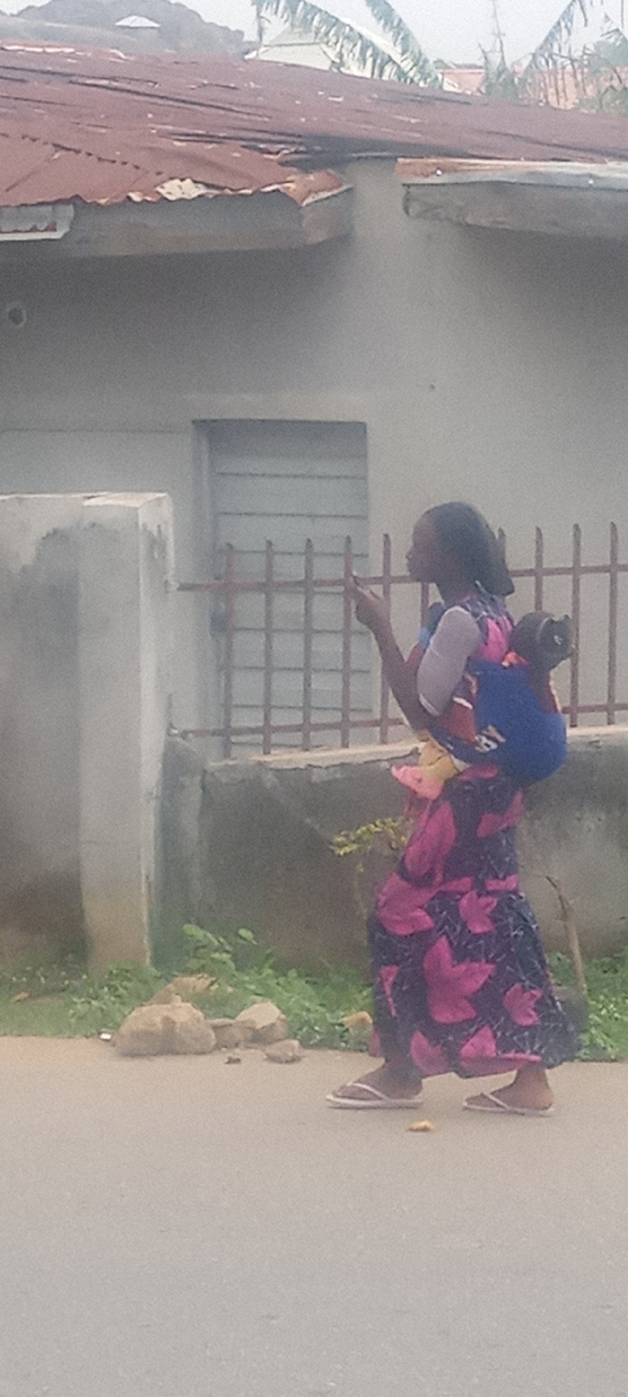 A woman carries a child on her back while walking on a street in Unknown, Unknown. The woman is wearing a long, floral-patterned dress, a light-colored top, and white flip-flops. She has a child secured on her back with a blue carrier. The woman is looking to her right and holding a small object in her right hand. Behind her is a building with a rusty corrugated metal roof and a white wall. The building has a barred window. There is a short concrete wall in front of the building with some vegetation growing along it. A portion of the street is visible in the foreground.
