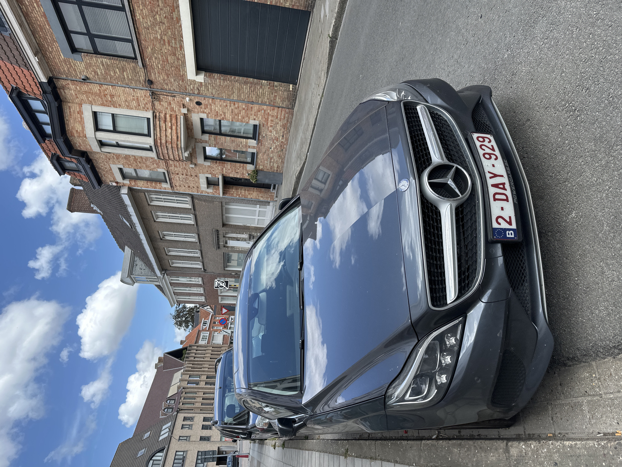 A dark gray Mercedes-Benz CLS is parked on a narrow, paved street in Bruges, Belgium. The car faces the viewer, angled slightly to the right. Its license plate reads "2-DAY-929". Adjacent to the car is a concrete sidewalk with some cigarette butts. To the right of the car, a brick building with a dark gray garage door is visible. Further down the street, other residential buildings with traditional Flemish architecture can be seen. The sky is blue with scattered white clouds. The image appears to be taken during daylight.