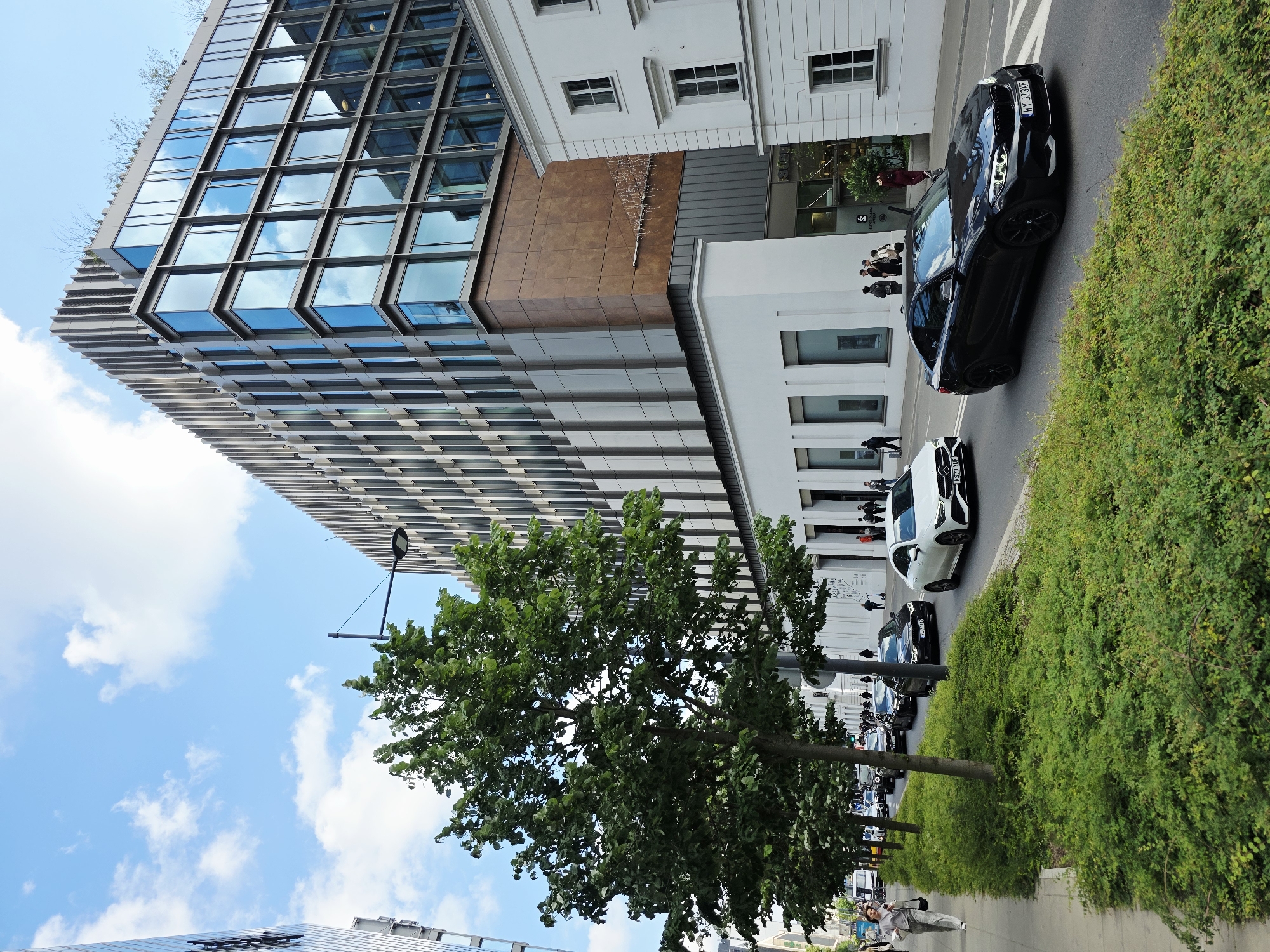 In Warsaw, Poland, a street scene unfolds with a modern glass and metal office building dominating the background. The building features a distinctive bronze-colored section and a ribbed facade. In front of it, a row of green trees lines the sidewalk, which is populated by several pedestrians. A street with moderate car traffic runs alongside the trees and sidewalk. Cars visible include a white Mercedes-Benz sedan and a black BMW coupe, both with visible license plates. The sky is blue with scattered white clouds, indicating daytime. The environment appears to be an urban setting with well-maintained landscaping.