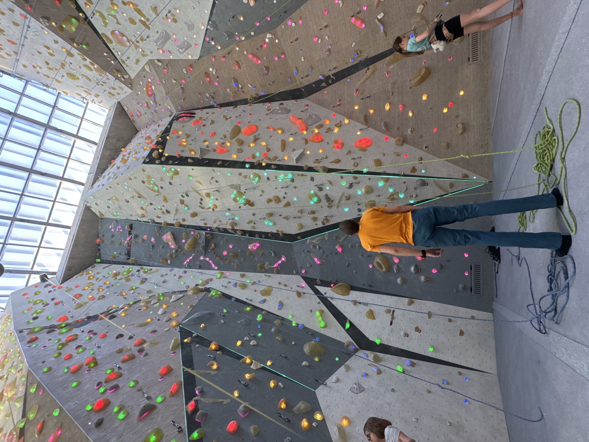 This image captures a dynamic scene inside a modern indoor climbing gym in Sofia, Bulgaria, characterized by its brightly colored, illuminated holds and geometric wall structures. The perspective is tilted, looking upwards at the climbing walls, making the floor appear as a vertical surface.
In the center-right of the frame, a person wearing an orange t-shirt and blue trousers is positioned on the floor, actively managing a yellow-green climbing rope. This individual is likely belaying. Above and to the left of them, a young person, possibly a girl or teenager, is actively climbing the textured wall. She wears a light blue t-shirt and black shorts with a climbing harness, and notably, is barefoot.
The climbing walls themselves are a striking feature, constructed with various shades of grey panels and delineated by black lines. They are densely populated with numerous climbing holds of diverse shapes and sizes, illuminated in vivid colors including red, green, blue, yellow, pink, and clear/white, suggesting a sophisticated route-setting system or interactive holds. A large skylight or window structure at the top-left provides natural light to the expansive space. Another person in a white shirt is partially visible at the bottom-right, near a coiled blue climbing rope. No discernible text is visible in the image.