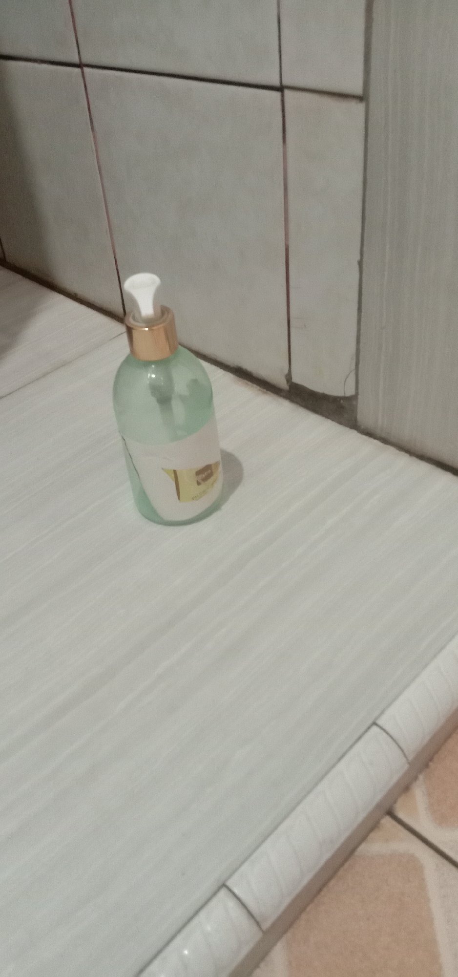 A clear plastic bottle with a pump dispenser is placed on a tiled surface. The bottle contains a light green liquid. A label, mostly obscured, is attached to the bottle. The background consists of light-colored tiles. The lighting appears dim, suggesting an indoor environment, possibly a bathroom or kitchen area. The image is taken from a slightly elevated angle, looking down at the bottle. The floor tiles are light grey with a subtle striped pattern. The wall tiles are also light-colored, arranged in a grid pattern with darker grout lines. The overall scene is clean and orderly, with no other objects or individuals present. This image does not provide any information that would allow for its location to be identified as Kendari, Indonesia.