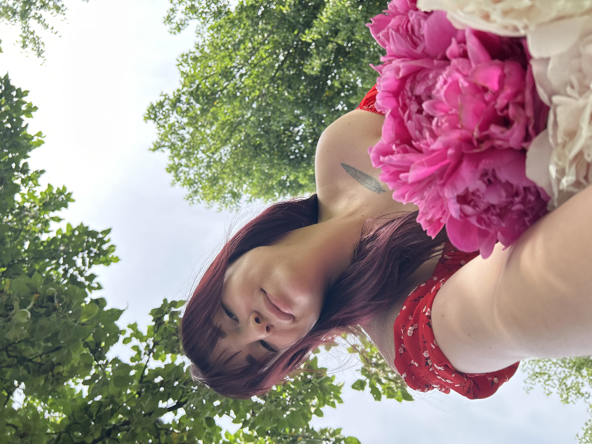 The image shows a young woman with long, dark red hair and bangs, wearing a red off-the-shoulder dress with a floral pattern. She is holding a bouquet of bright pink and white peonies. She is looking up and smiling slightly, with her head tilted. Her left shoulder is visible, and she has a tattoo of a feather on her upper arm. The background consists of lush green trees and a bright, overcast sky. The photo appears to be taken outdoors, possibly in a park or garden. The angle of the shot is from below, looking up at the woman. The location of Фоки, Russia is not discernible from the image.