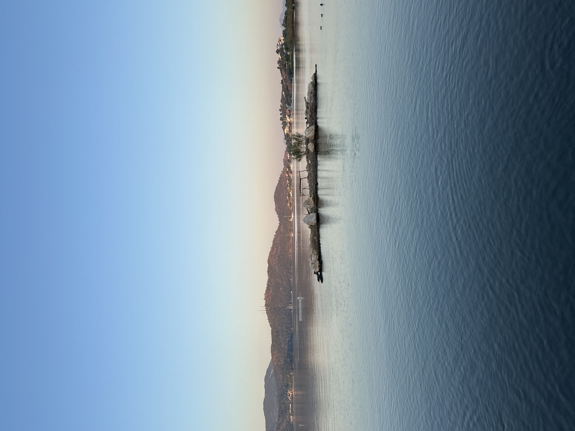 A wide shot captures a serene body of water in Fethiye, Turkey, at what appears to be sunrise or sunset. The sky is a clear, soft gradient of pale blue to light pink near the horizon. Across the water, a line of low, tree-covered hills stretches into the distance, dotted with buildings and communication towers on the highest peak. In the foreground, a small, rocky islet hosts a sparse clump of vegetation and some driftwood. Several birds are visible in the water to the right of the islet. The water itself is calm, with subtle ripples reflecting the soft light, creating a peaceful and expansive atmosphere. The overall scene is tranquil and natural.