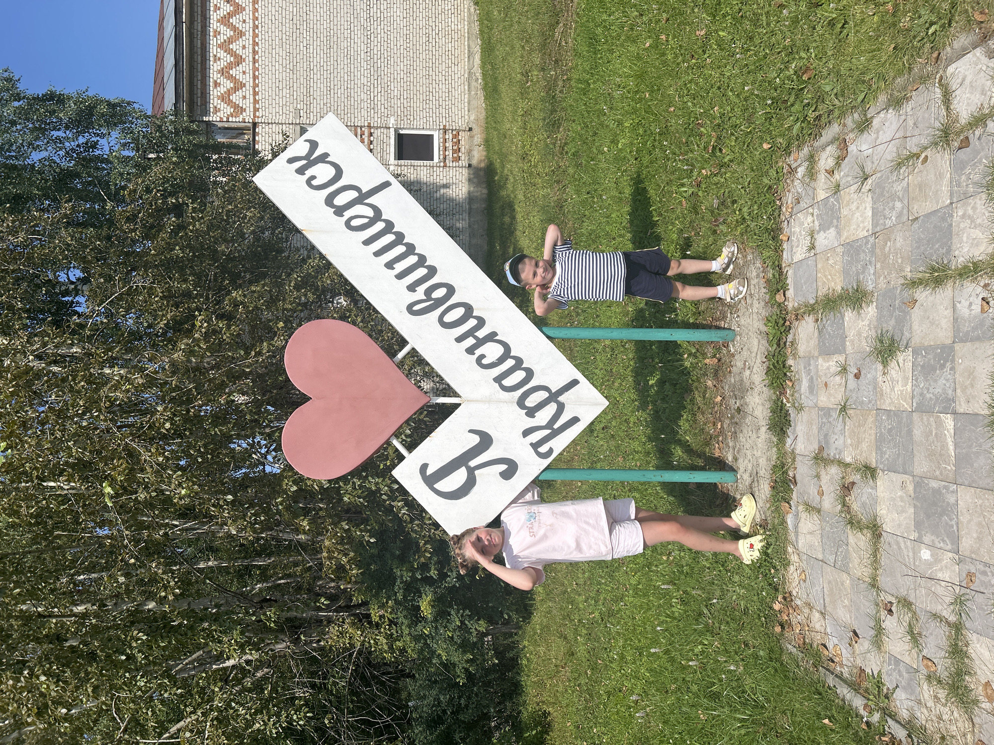 In Krasnovishersk, Russia, a large sign with a pink heart and white text that reads "Я люблю Красновишерск" (I love Krasnovishersk) stands in a grassy area with trees and a brick building in the background. Two children are posing in front of the sign. On the left, a young girl with blonde hair in pigtails wears a pink t-shirt and white shorts. On the right, a young boy wearing a striped t-shirt and dark shorts with a blue cap strikes a playful pose. The ground in front of the sign is paved with square tiles.