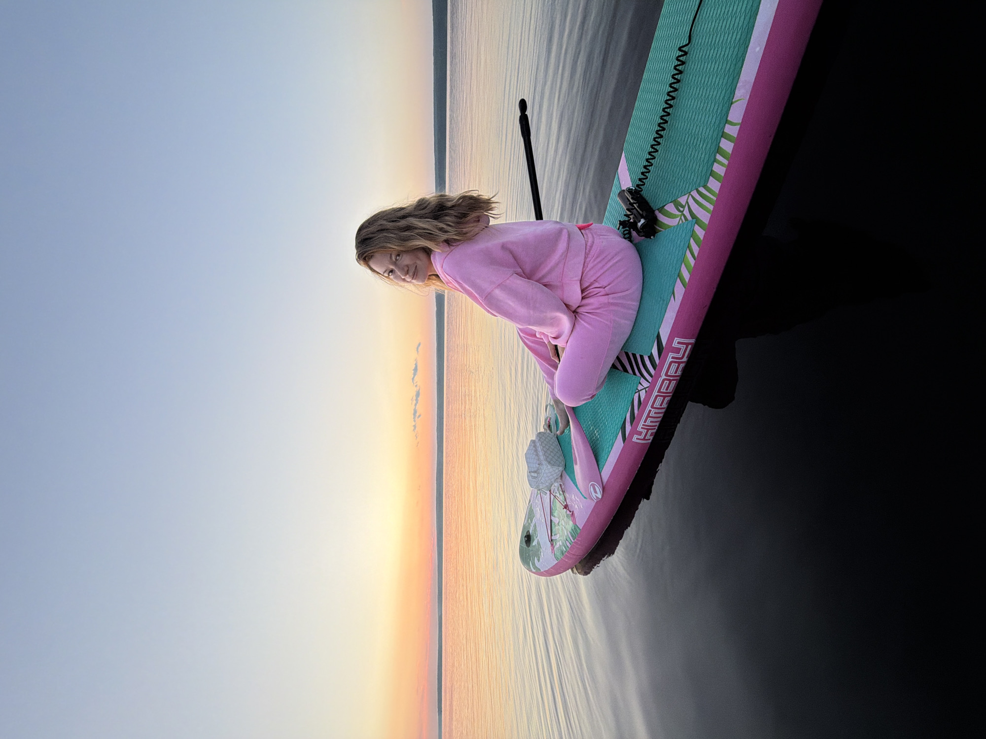 A young woman with long, blonde hair sits cross-legged on a paddleboard in a calm body of water. She is wearing a pink tracksuit and a white handbag is beside her. The paddleboard is pink and teal with a palm leaf pattern. The sky is a gradient of soft blues and oranges, indicating sunset or sunrise. The water is dark and reflective, with the paddleboard casting a shadow. The distant horizon shows a line of trees. The scene appears to be set on the Kama River in the city of Chaikovsky, Russia, known for its scenic waterfront and recreational activities.