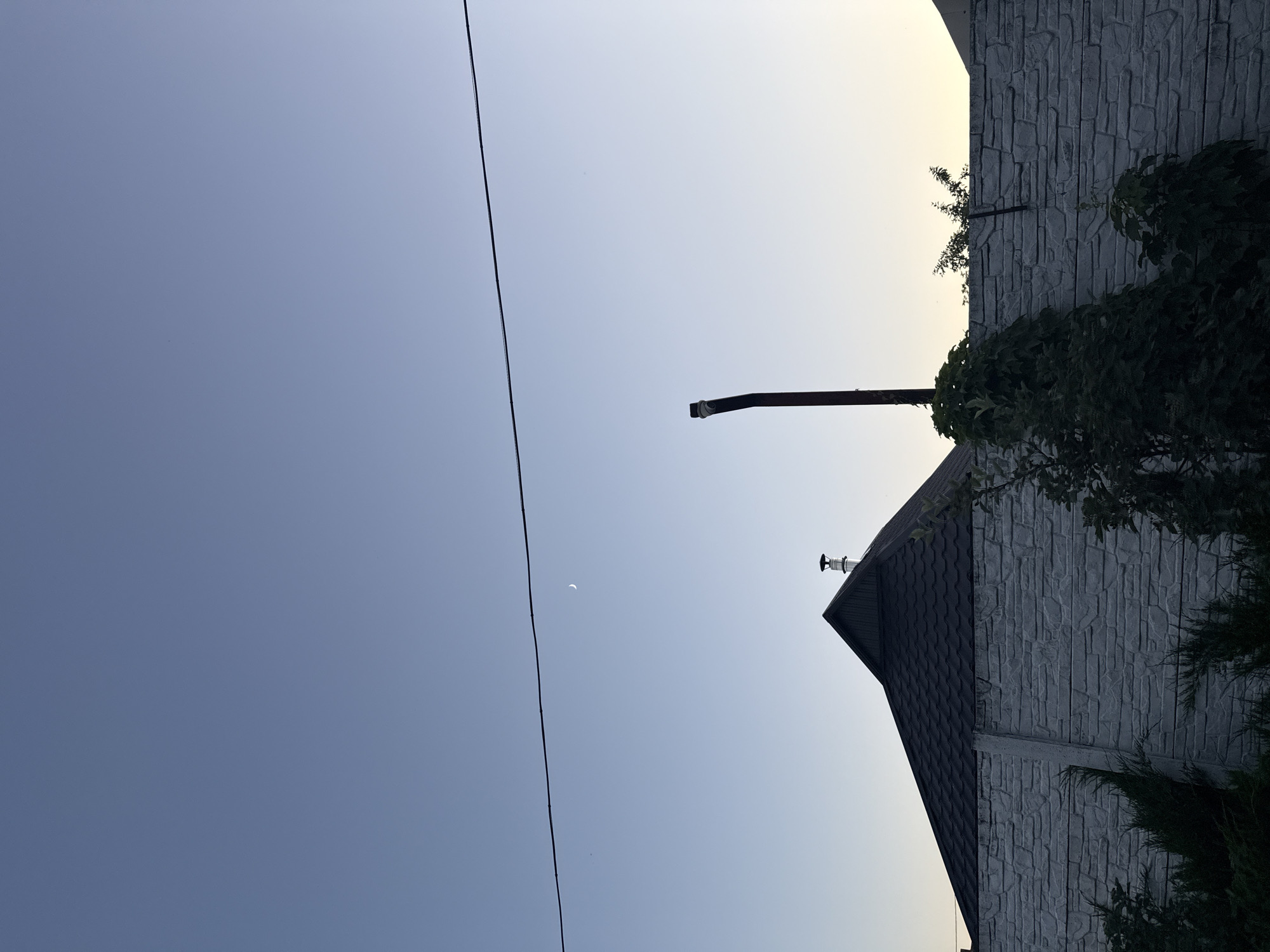 In Obukhivka, Ukraine, at dusk, a crescent moon is visible in the clear, light blue sky. A thin black power line stretches horizontally across the upper portion of the frame. Below the sky, a dark-pitched roof of a house with a chimney is silhouetted against the lighter horizon. A textured, light gray concrete fence runs across the middle ground, partially obscuring greenery. A dark pipe, possibly a vent or chimney extension, rises vertically from behind the fence on the right side.