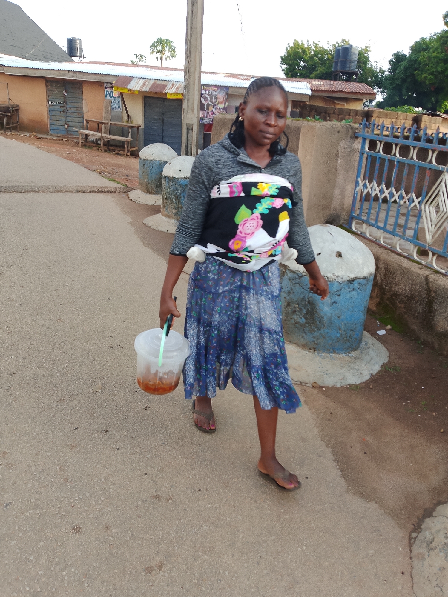 A Nigerian woman is walking on a paved street in Jos, Nigeria, carrying a transparent plastic bucket containing a dark liquid. She is wearing a grey long-sleeved shirt, a colorful patterned wrap around her midsection, and a blue floral print skirt. Her hair is braided and pulled back. In her right hand, she holds the bucket by its handle. She is wearing flip-flops. The environment consists of buildings with corrugated metal roofs, a blue metal gate, and concrete bollards along the side of the street. The sky is overcast.