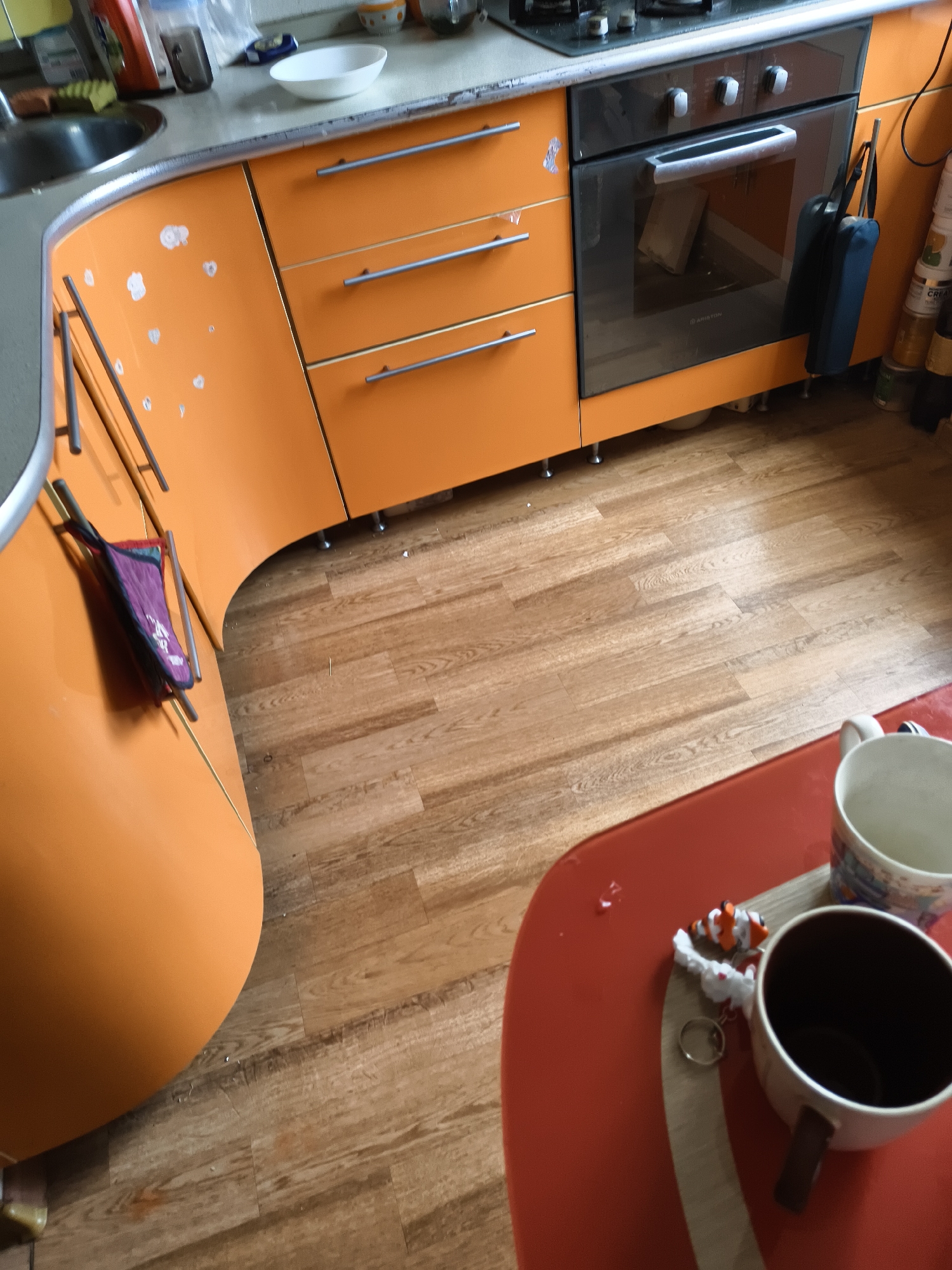 The image shows a close-up, high-angle view of an orange kitchen with light wood laminate flooring. In the foreground, a red table has two mugs and a clownfish keychain. In the middle ground, there is an orange kitchen cabinet with drawers and an oven. In the background, there are various kitchen items on a countertop, including a sink and a white bowl. The scene appears to be indoors, possibly in a residential kitchen. There are no people visible in the image.