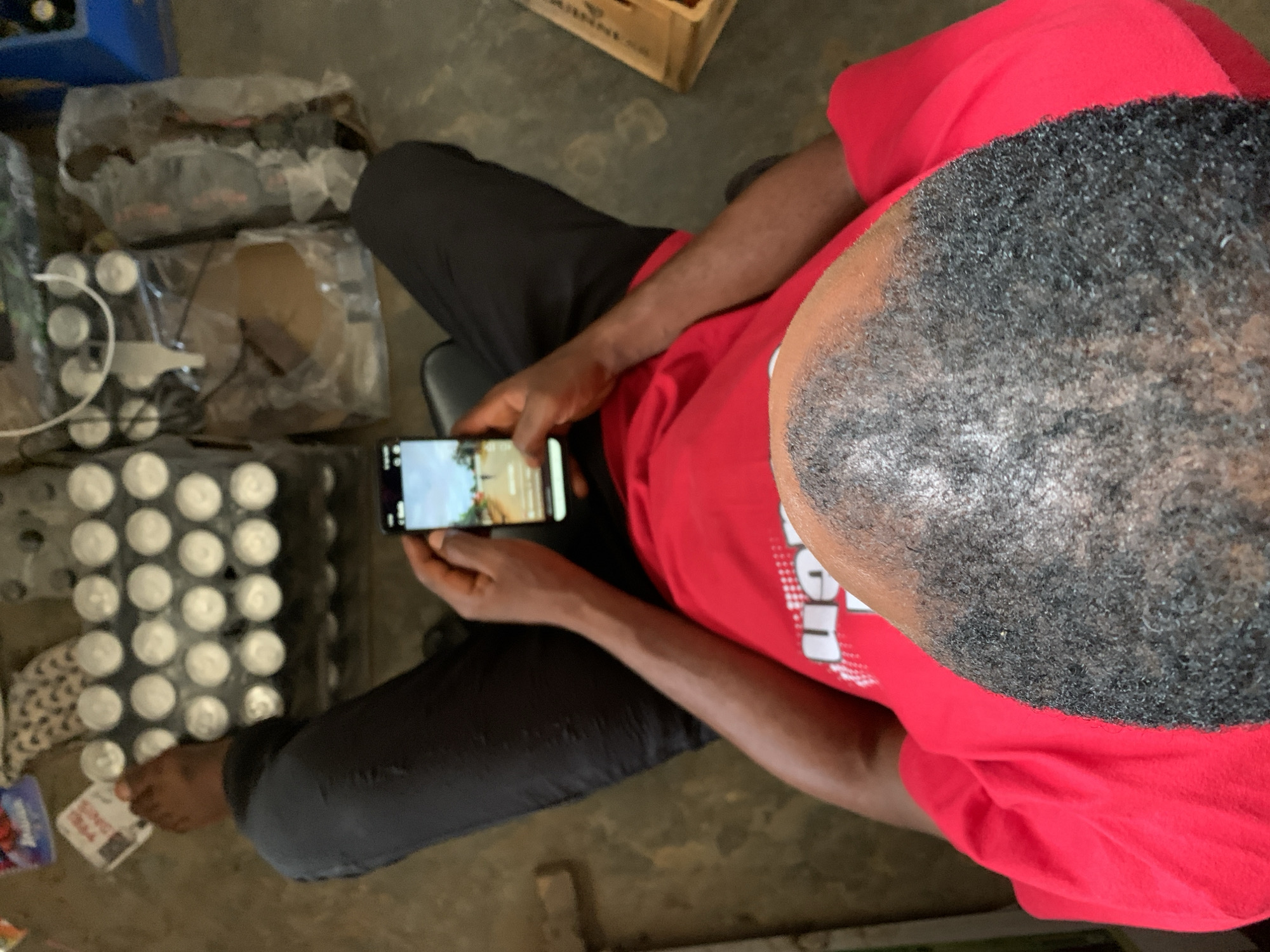 The image shows a person sitting on a chair, looking at their phone. The person is wearing a red shirt and black pants. Around them, there are several packs of cans, some wrapped in plastic, and a wooden crate in the background. The location is identified as New Stadium, Nigeria. The person is holding the phone horizontally and seems to be viewing content on the screen. One bare foot is visible near the cans. The environment appears to be an indoor space, possibly a storage area or a small shop.