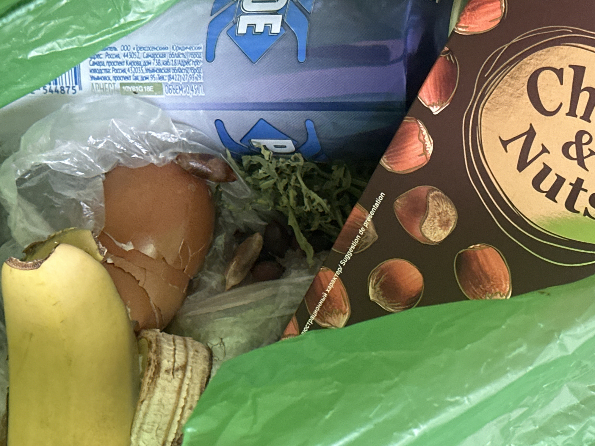 The image shows the inside of a green plastic bag. A partially peeled banana and a broken eggshell are visible. There is also a blue and silver can with the word "DE" visible on it. A portion of a chocolate box with images of hazelnuts is also present. The text on the chocolate box appears to be in Russian and English, stating "Иллюстрационный характер/ Suggestion de présenta". There is also some green leafy material in the bag. The scene is likely a trash bag, given the presence of food waste and packaging. No people or animals are present. The location is not specified within the image.