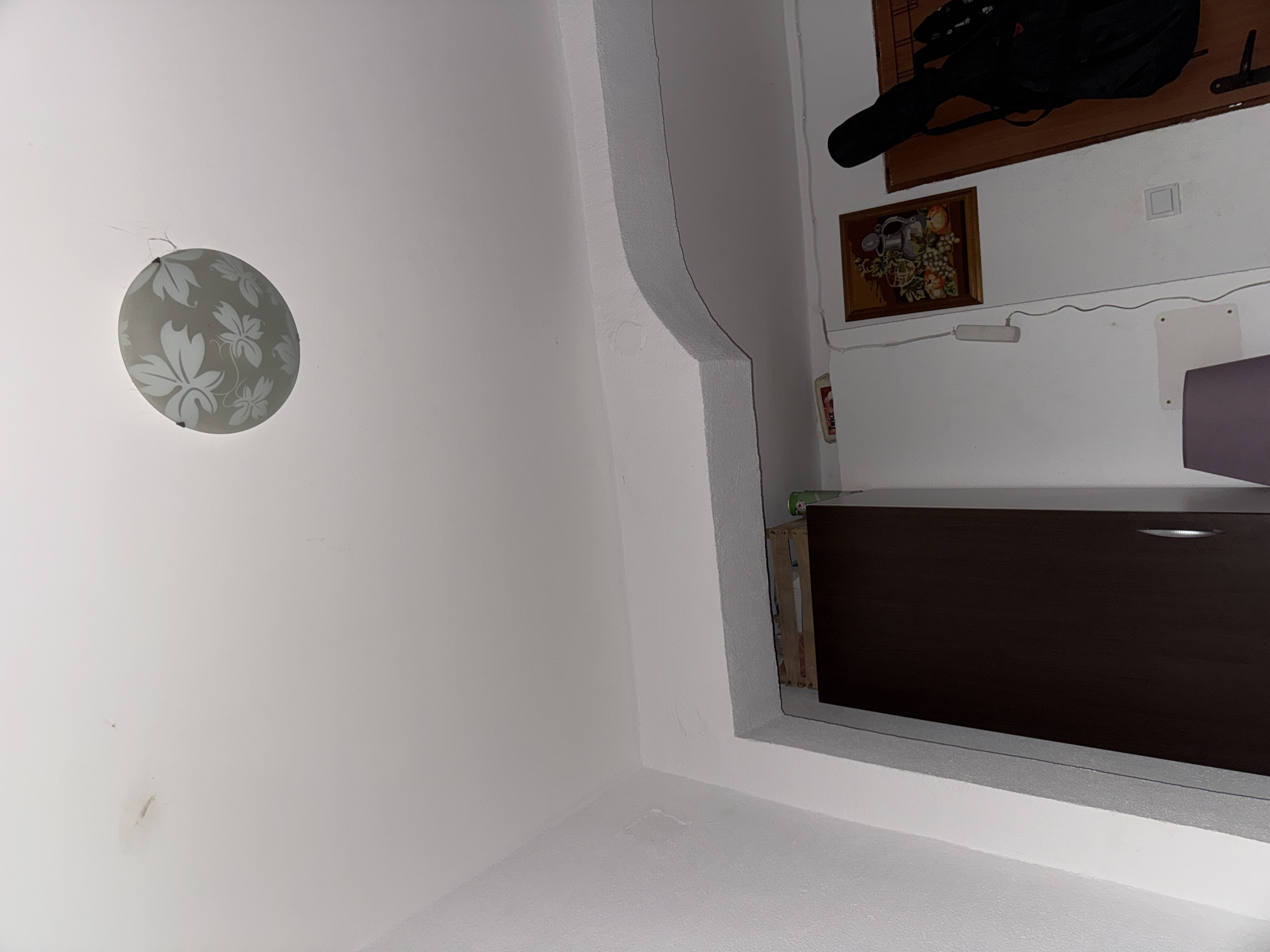 This is a medium-shot, indoor photograph taken from a low angle looking upwards. The scene appears to be a room in a residence in Sutomore, Montenegro. The ceiling is white and textured, with a decorative light fixture featuring a frosted glass shade with a floral pattern. The wall to the left is also white and textured. In the center of the image, a dark brown wardrobe is partially visible. Above the wardrobe, there is an arched architectural feature in the ceiling. On the wall to the right, a framed tapestry depicting a still life of fruit and a pitcher hangs. To the right of the tapestry, a dark-colored guitar case is propped upright. Several electrical cords are visible running along the wall. A white light switch is also present on the wall. The overall impression is of a somewhat cluttered living space.