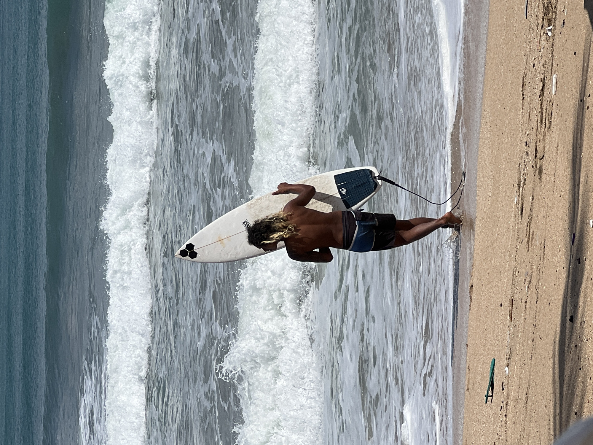 In Kuta, Indonesia, a young surfer with blonde, curly hair stands with his back to the viewer, facing the ocean. He is holding a white surfboard vertically in his right hand. The surfer is wearing dark swim trunks. He is standing in the shallow water where gentle waves are breaking. The beach in the foreground is sandy and appears to have some debris. The ocean is a deep blue with white foam from the waves stretching to the horizon.