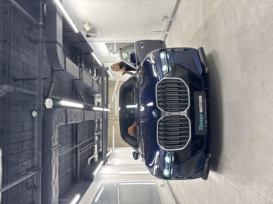 The media features a dark blue BMW car, likely a sedan or SUV, parked in an indoor setting that resembles an industrial-chic showroom or garage in Warsaw, Poland. The car, positioned towards the center-right, has a prominent large kidney grille and sleek headlights. On its front license plate holder, the text "TIFFANY MOTORS" is clearly visible, with "DYNAMIC MOTORS GROUP" and small M logos below it.
A woman with blonde hair is partially visible, leaning into the open driver's side door of the car, seemingly interacting with the interior. The car's interior features tan or light brown upholstery, visible through the open door.
The background on the left consists of a dark grey wall with a brick-like texture. This wall is adorned with extensive exposed black and grey utility infrastructure, including large ducts, pipes, and various fixtures, some of which appear insulated. Bright fluorescent strip lights are mounted on the wall and potentially the ceiling, illuminating the scene. The floor is a light grey concrete, and the ceiling/upper wall area is white. The overall setting combines a modern luxury car with an raw, industrial aesthetic.