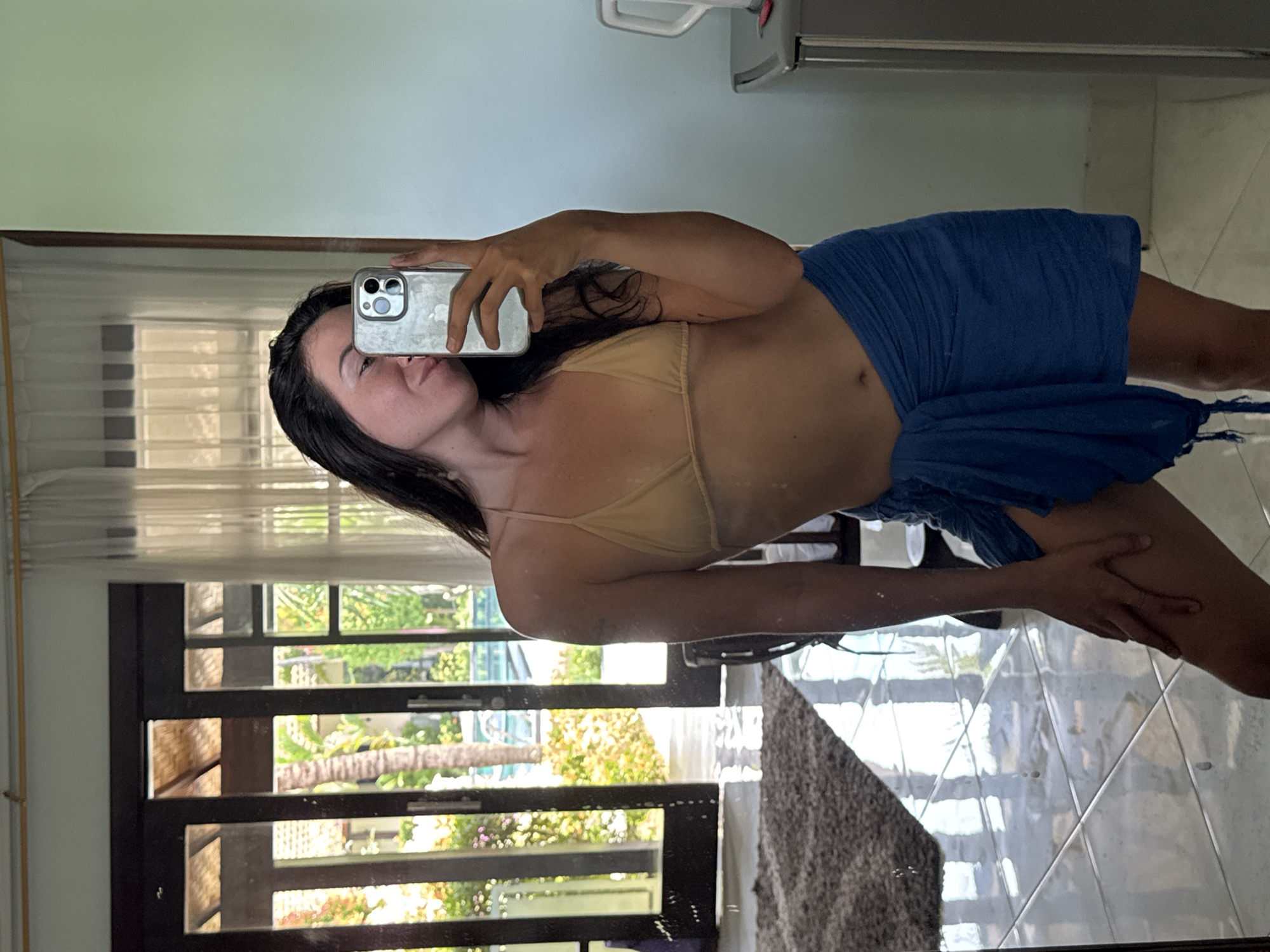 A young woman with dark hair takes a mirror selfie in a room that appears to be within a hotel or resort in Karangasem, Indonesia. She is wearing a light tan bikini top and a bright blue sarong tied around her waist. Her left hand holds up a smartphone to capture the reflection. Through a set of open glass doors behind her, a sunlit outdoor area is visible, featuring palm trees and what appears to be a swimming pool. The floor is tiled and reflects the light from the outdoor area and the room. The room itself is sparsely furnished, with a glimpse of a plush rug and a cabinet with a kettle on top to the right.