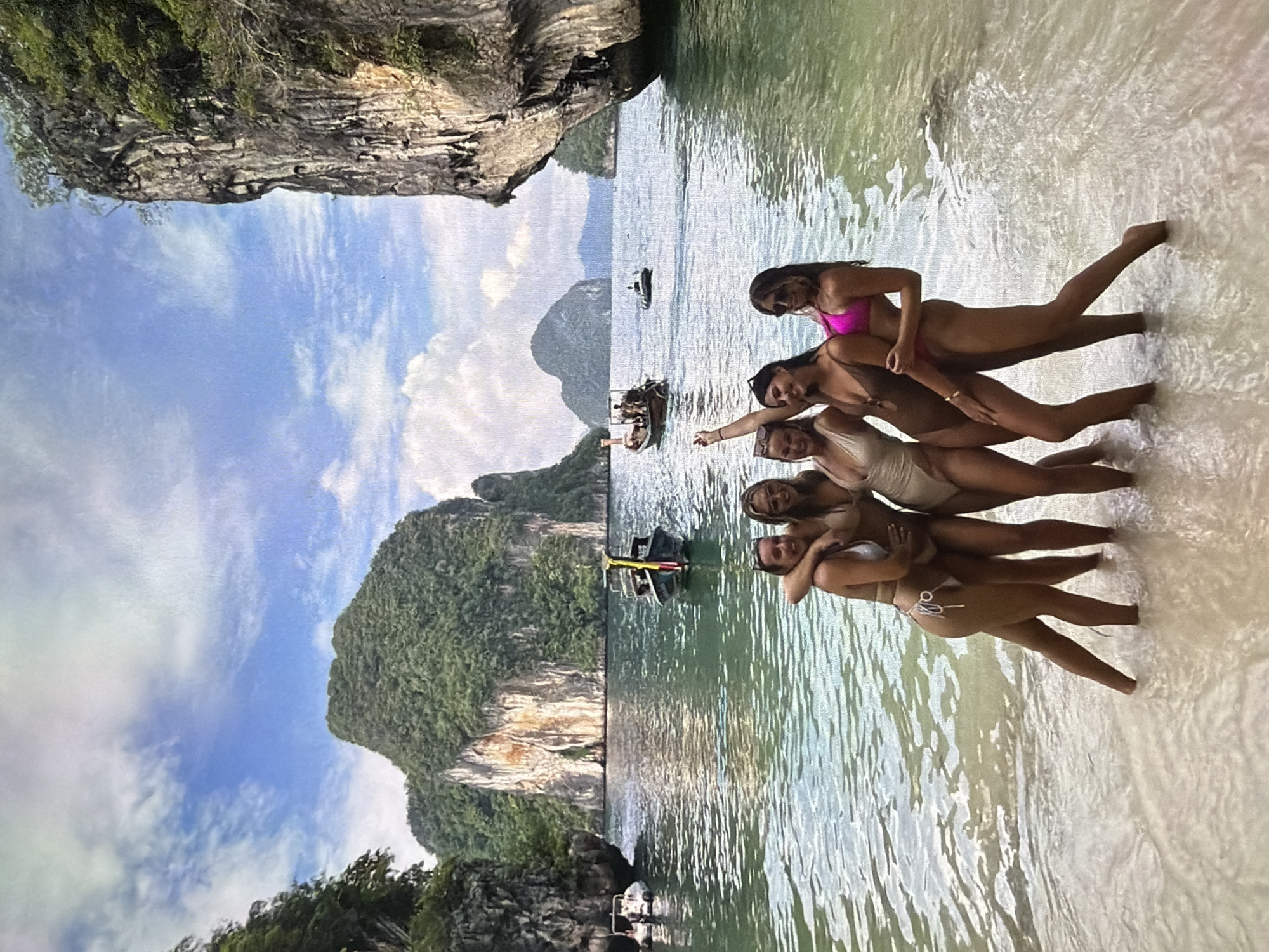 The image captures a group of six women posing in shallow, clear turquoise water at a scenic tropical beach. The women, all wearing swimsuits (including pink, tan, and white), are arranged in a playful, stacked formation, with the woman closest to the viewer embracing another. They appear to be enjoying their time, with relaxed postures and smiling expressions.
The background features majestic limestone karst islands, characteristic of Southeast Asian coastlines, such as those found in Thailand or Vietnam. These dramatic cliffs are covered in lush green vegetation. The water transitions from a light sandy color near the shore to a deeper blue-green further out, where several boats, including what resemble longtail boats, are visible. The sky is bright blue with scattered white clouds, indicating sunny weather during the daytime. The overall atmosphere is one of leisure and vacation. There is no visible text in the image.
