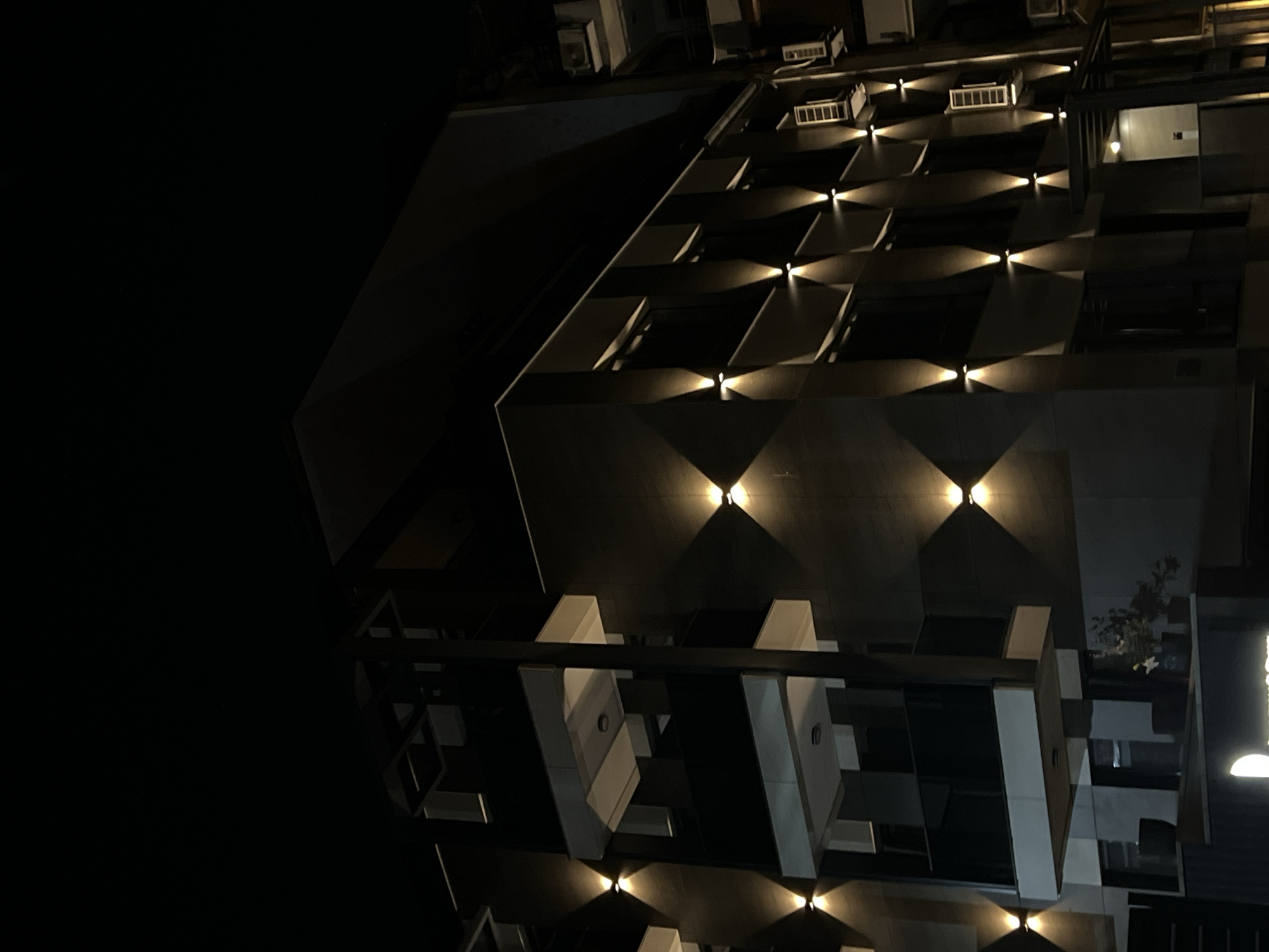 The image depicts a modern building at night, illuminated by strategically placed wall lights that create diamond-shaped patterns of light and shadow on its facade. The building appears to be multi-story, with visible balconies and windows. Air conditioning units are mounted on the exterior walls. The scene suggests an urban setting. The specific location in the city of Sofia, Bulgaria, is not discernable from the visual information alone. There are no people visible in the image. The lighting creates a dramatic and somewhat abstract aesthetic.