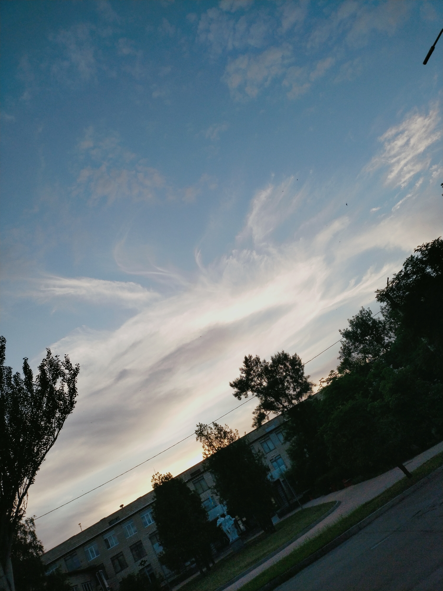 The image depicts an outdoor scene, likely in Horishni Plavni, Ukraine, during what appears to be late afternoon or early evening. The sky is a prominent feature, filled with a soft, pale blue color and streaked with delicate, wispy white clouds, suggesting fair weather. A faint glow on the horizon indicates the setting sun, casting a warm light that subtly illuminates the clouds.
In the foreground and middle ground, a building is visible, constructed from light-colored bricks and featuring multiple windows, some of which appear to have blue-tinted panes. The building is angled diagonally across the frame, running from the lower left to the upper right. Trees, mostly dark green and silhouetted against the brighter sky, line the front of the building and extend into the background, creating a sense of depth. Some of these trees are tall and leafy, while others are more sparse.
A paved area, possibly a road or walkway, runs along the bottom right of the image. The overall atmosphere is peaceful and serene, with the natural beauty of the sky contrasting with the more man-made elements of the building and paved surfaces. There are no people or explicit activities depicted. A thin, dark line, possibly a power line, stretches across the frame from left to right, intersecting the building and trees. A tall, dark, rod-like object, possibly a lamp post or antenna, is visible in the upper right corner of the sky. No text is discernible in the image.