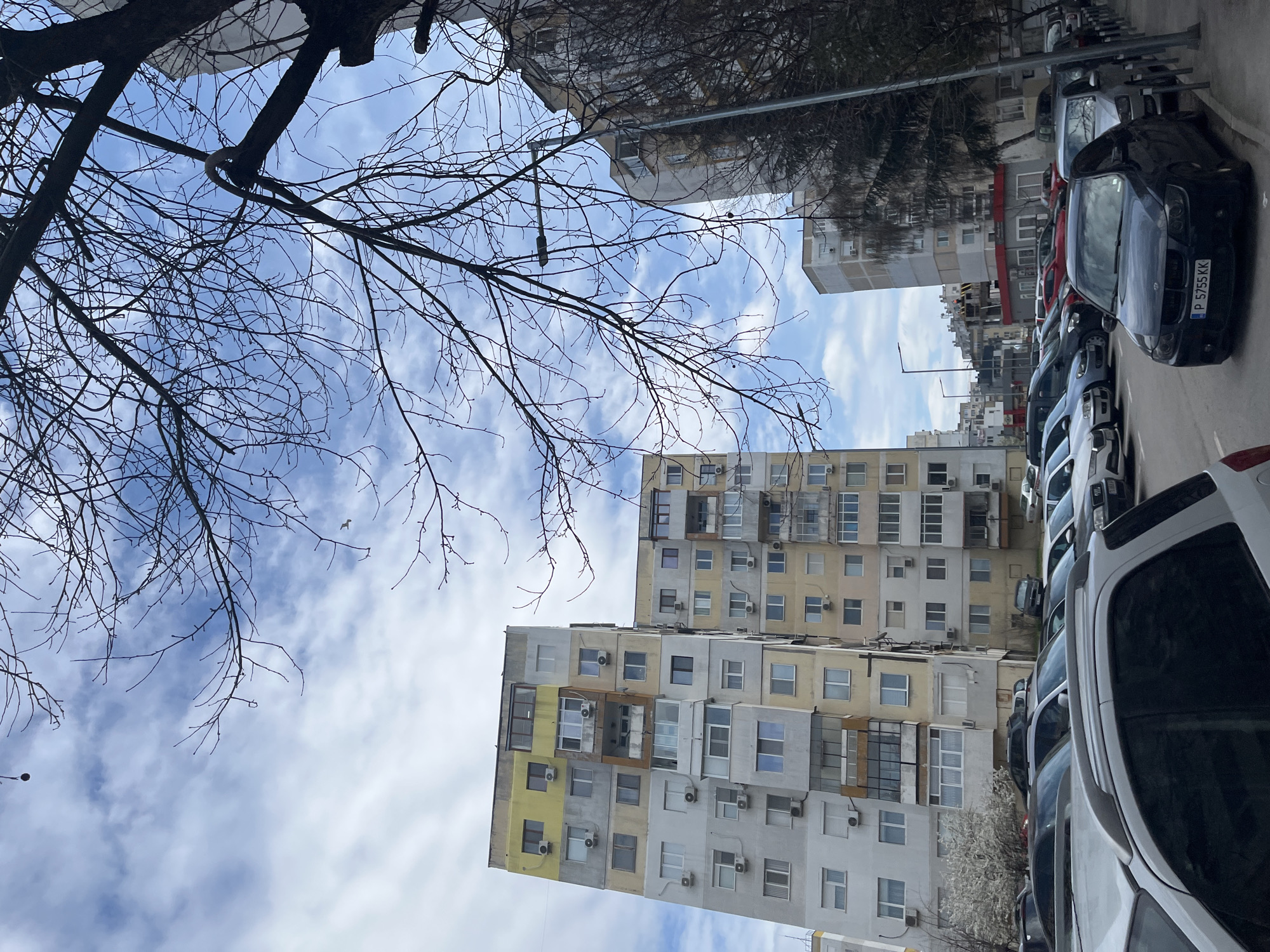 A street scene in Varna, Bulgaria. The photograph is taken looking upwards at an angle, capturing a row of parked cars on the right side of the frame. Several high-rise residential buildings stand in the background, with varying colors and architectural styles, with many of the buildings having visible air conditioning units. Bare tree branches are visible in the upper left quadrant, set against a sky with white clouds. The photograph appears to be taken on a partly sunny day.