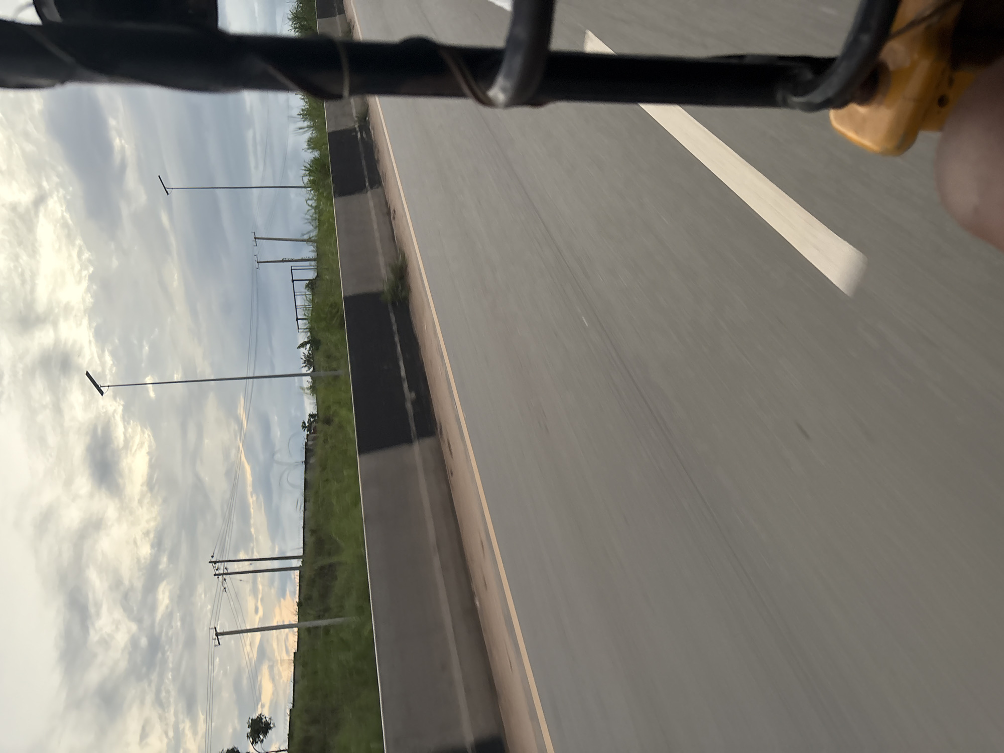 The image captures a view from a moving vehicle, likely a tuk-tuk, on a road in Onitsha, Nigeria. The foreground shows a portion of the vehicle's structure and a passenger's leg. The road ahead is asphalt with a clear white line marking a lane. A concrete barrier, painted with black and grey sections, separates the road from a grassy embankment. Beyond the embankment, utility poles with power lines and modern streetlights are visible against a cloudy sky. The scene suggests a typical urban or suburban environment with ongoing infrastructure.