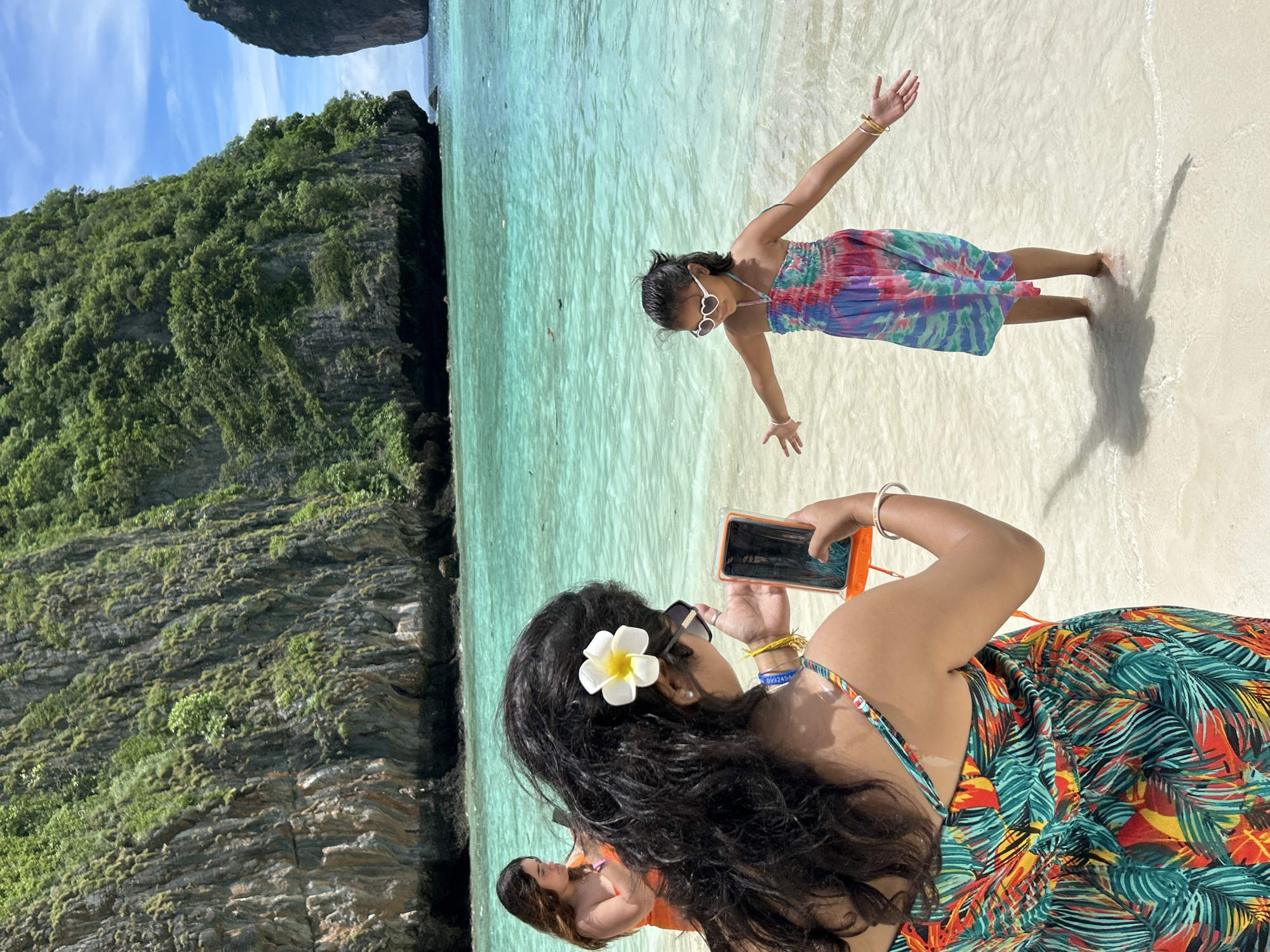 In this vertical, eye-level, medium shot, two individuals are captured on a sunlit beach, with a turquoise ocean and lush green cliffs in the background. The scene appears to be Ao Nang, Thailand. A young girl in a colorful tie-dye dress and heart-shaped sunglasses is standing in the shallow water with her arms outstretched, her face turned towards the camera. Beside her, partially in the foreground, a woman with dark, wavy hair adorned with a white and yellow flower is taking a photo with her orange-cased smartphone. She wears a vibrant tropical-print dress. A second woman, wearing an orange swimsuit, is visible in the lower left corner, partially obscured. The sun is bright, casting shadows on the sand and water. The overall mood is joyous and relaxed, indicative of a vacation. The image is well-lit, suggesting it was taken during daylight hours. No visible text is present in the image.