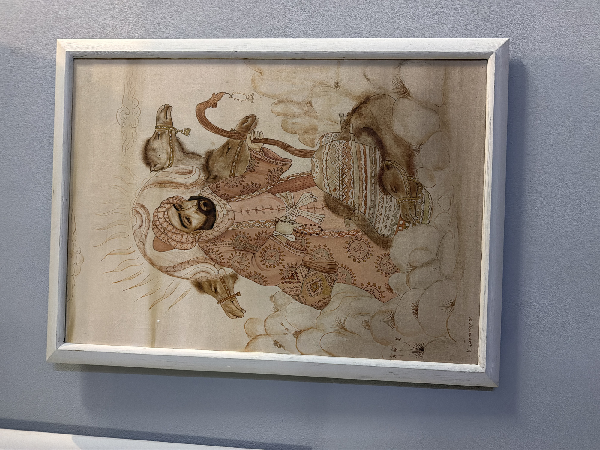 A framed painting depicts a man in traditional attire with a beard and turban, holding a staff and standing amidst camels in a desert landscape. The man wears a pink robe adorned with intricate patterns. Two camels are visible, with their heads facing towards the man, and a third camel's head is partially obscured. They are situated on rocky terrain. In the background, a sun-like halo emanates from behind the man's head. The painting is signed "K. Ulugbek. 03" in the lower left corner. This artwork is located in the city of Chinarkent, Uzbekistan.