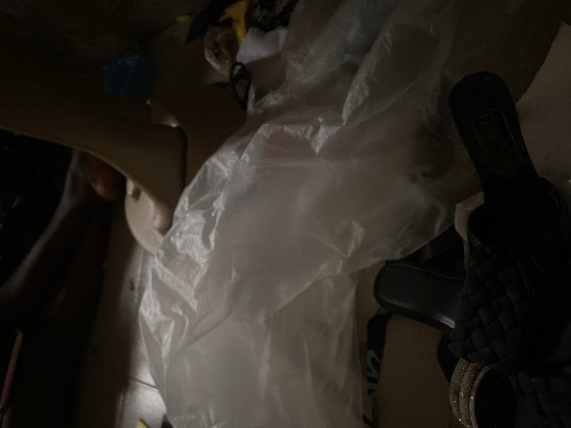The image depicts a close-up, low-angle view of discarded items on a tiled floor, likely within a residential setting in Jauro Yinu, Nigeria. In the foreground, a large, crumpled piece of translucent white plastic sheeting is prominent, obscuring much of the scene. To the left of the plastic, a black slide sandal with a woven texture is visible, partially covered by the sheeting. Next to it, another sandal or shoe is visible, with a portion of a light-colored, possibly sequined strap. Further to the right, the heel and sole of a black sandal with the embossed text "DIRECTOR SHOE" are visible. In the background, a human leg and foot, dressed in light-colored attire, are resting on the floor. The leg appears to be positioned above a small, blue, plastic-wrapped object. Other indistinct objects, possibly clothing or discarded materials, are scattered around the scene. The lighting is dim, suggesting either an indoor environment with low light or it's nighttime. The overall impression is one of a cluttered, unkempt area with discarded personal items.