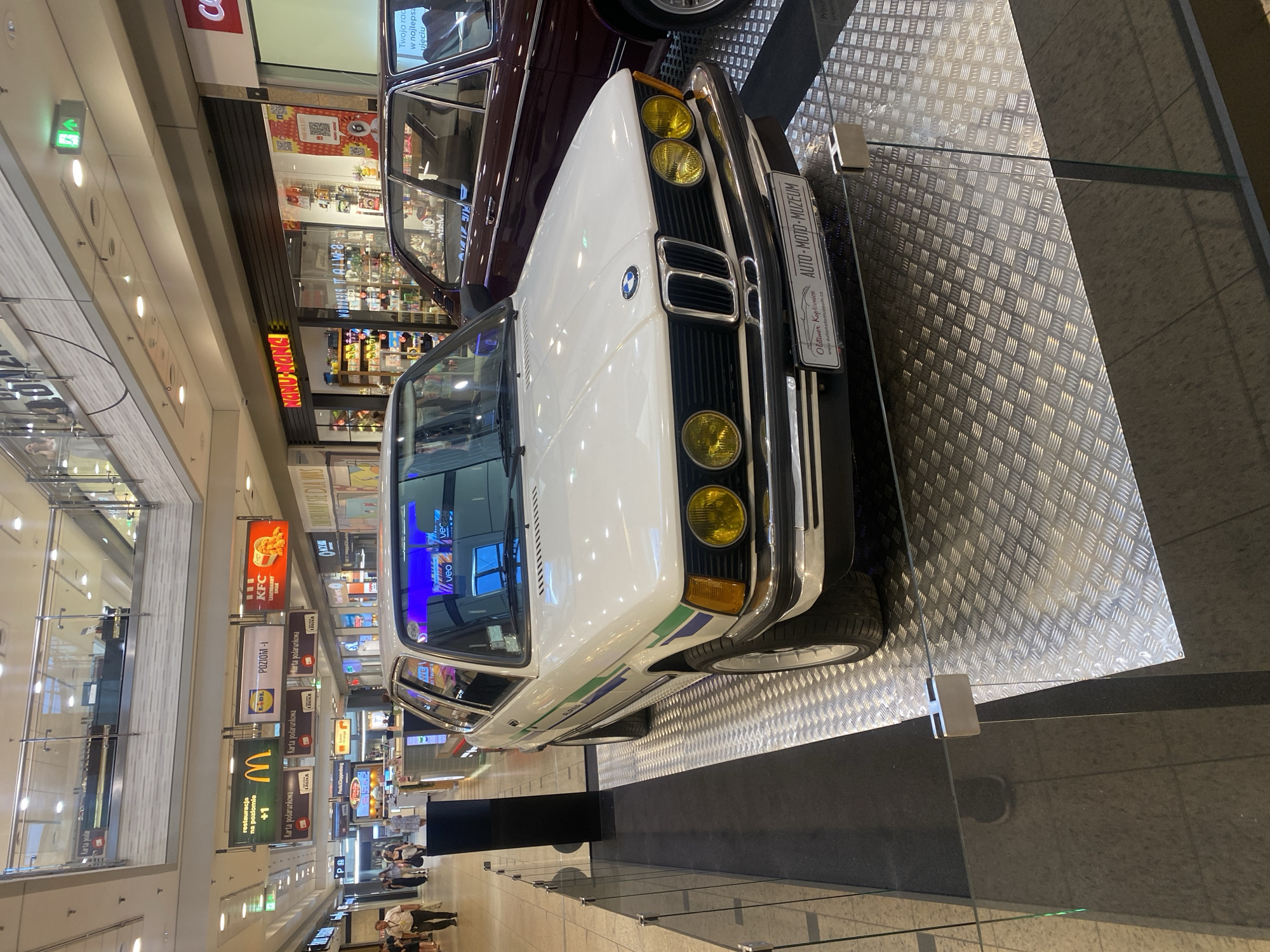 This media file shows a white classic BMW car displayed in a shopping mall in Łódź, Poland. The car is parked on a diamond plate platform, with a darker car parked behind it. Signs for various stores and restaurants are visible in the background, including KFC, Lidl, and McDonald's, as well as a sign for "AUTO-MOTO-MUZEUM" with the website "www.automotomuzeum.cz". There are a few people walking in the background, appearing to be shoppers. The lighting suggests it is daytime inside the mall. The car appears to be part of an exhibition or display.