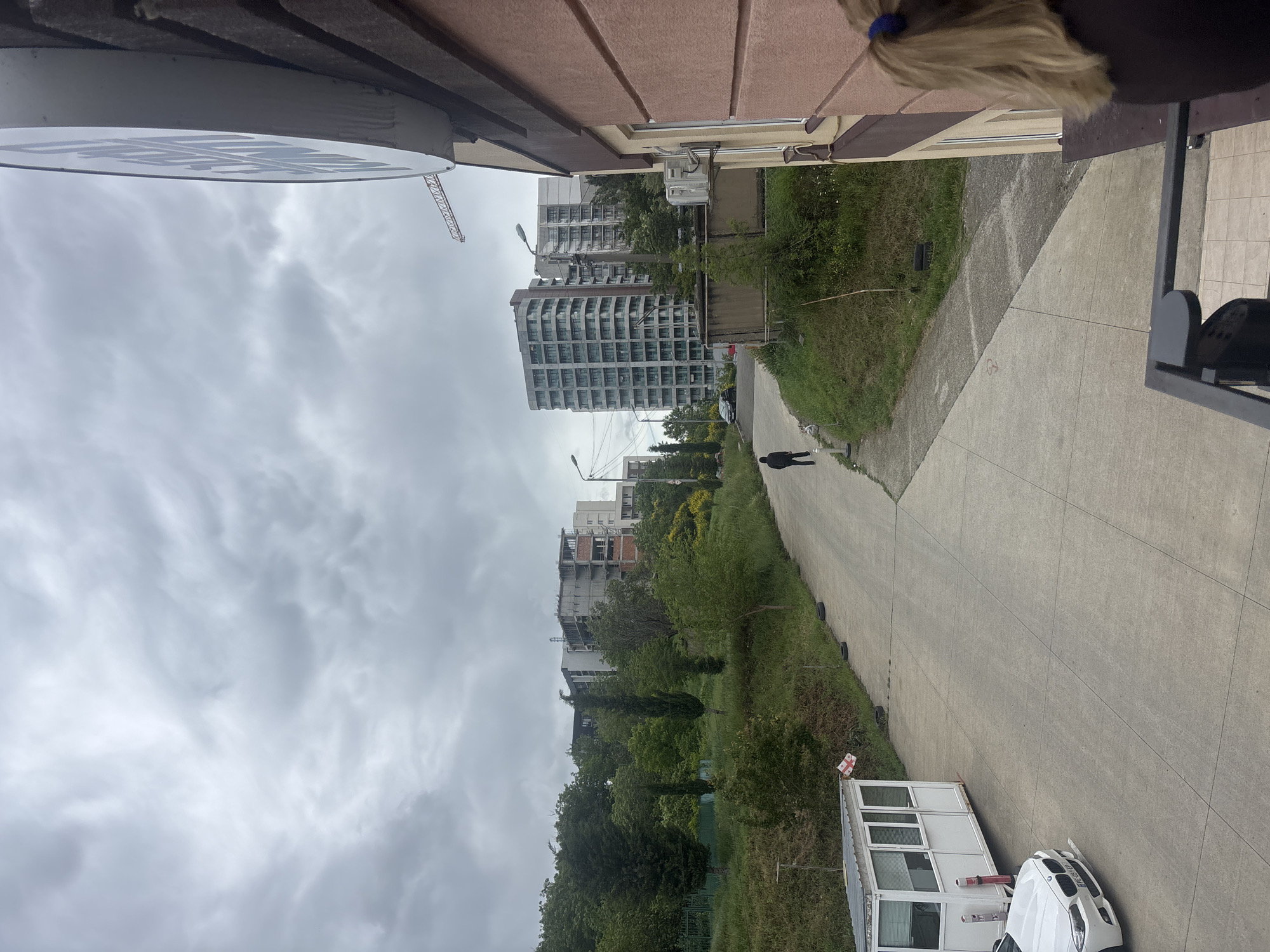 The image depicts an overcast view from an elevated position, likely a balcony, in what appears to be a cityscape in Tbilisi, Georgia. The scene is dominated by concrete structures, including a high-rise apartment building and a long, straight concrete path or road. A person is walking along this path. A white car is parked adjacent to the path near a small, white, portable building. Vegetation, including trees and grass, borders the path. The sky is heavily clouded. A construction crane is visible in the distance. The scene appears somewhat industrial or urban-fringe.