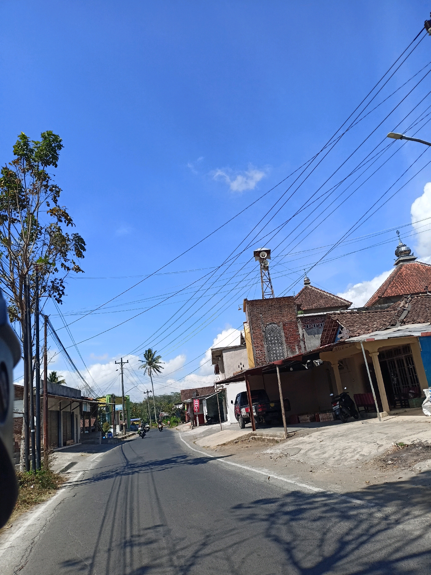The image depicts a street scene in Windusari, Indonesia. The road is paved and curves to the right, with a shoulder and sidewalks lining it. On the right side of the street, there are buildings with traditional tiled roofs, including what appears to be a mosque with minarets. Some buildings have open-fronted structures, possibly shops or garages, with motorcycles and a black pickup truck parked under them. There are also baskets of goods visible. On the left side of the street, there are more buildings, some appearing to be shops with rolled-down shutters. A tall tree with green leaves stands prominently on the left. The sky is a clear blue with a few scattered white clouds. Numerous power lines crisscross the sky, supported by utility poles. In the distance, more trees and buildings can be seen, suggesting a semi-urban or rural setting. A few motorcycles are visible on the road, with riders. The overall impression is of a sunny day in a residential and commercial area.